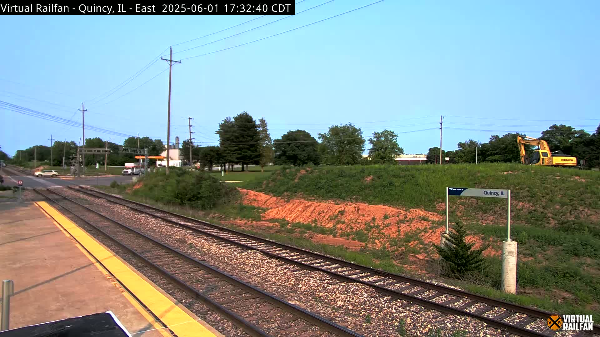 A train track and platform are shown, with a yellow excavator visible in the background under a clear blue sky.