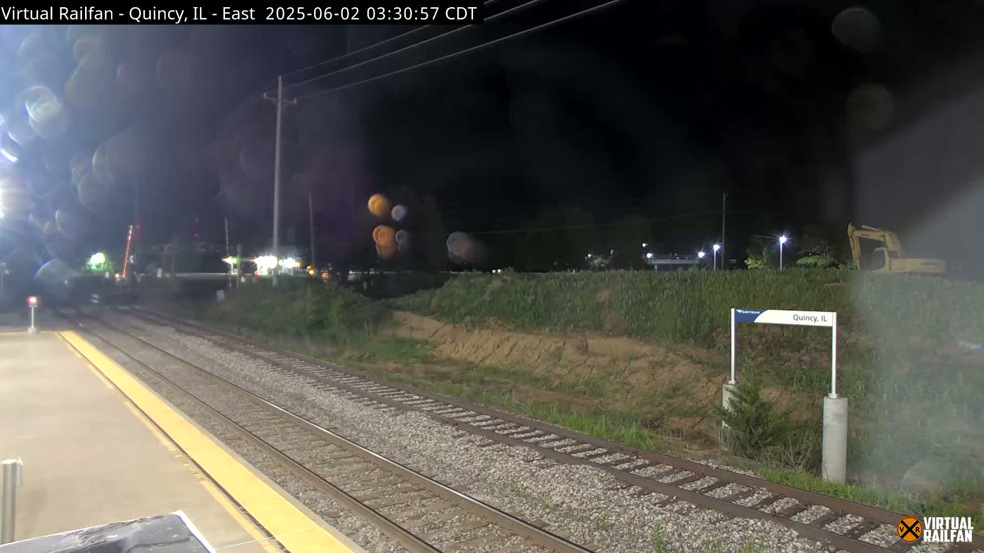 A nighttime view of a train station platform with train tracks, a small embankment with vegetation, and a piece of construction equipment in the background.