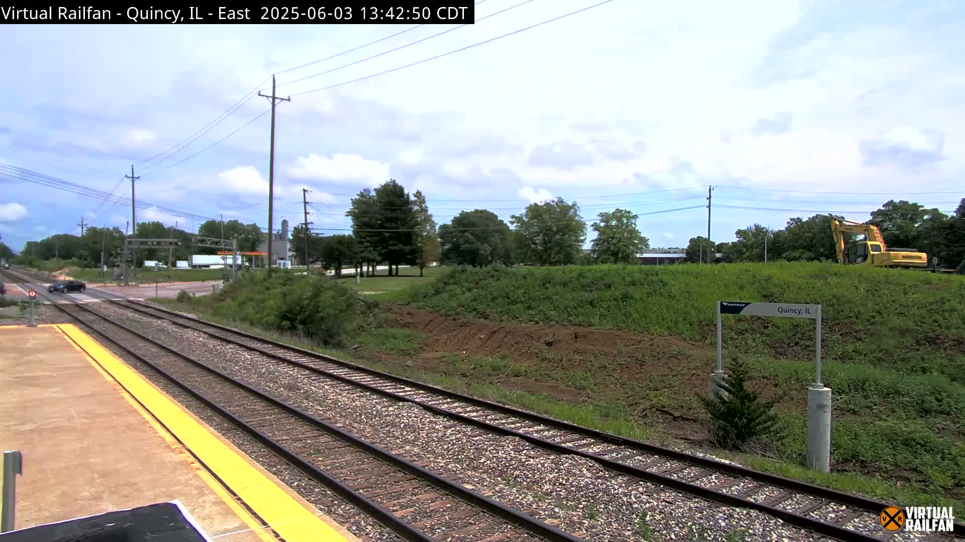 A partly cloudy day reveals a train platform, tracks, and a yellow excavator on an embankment beside a road.