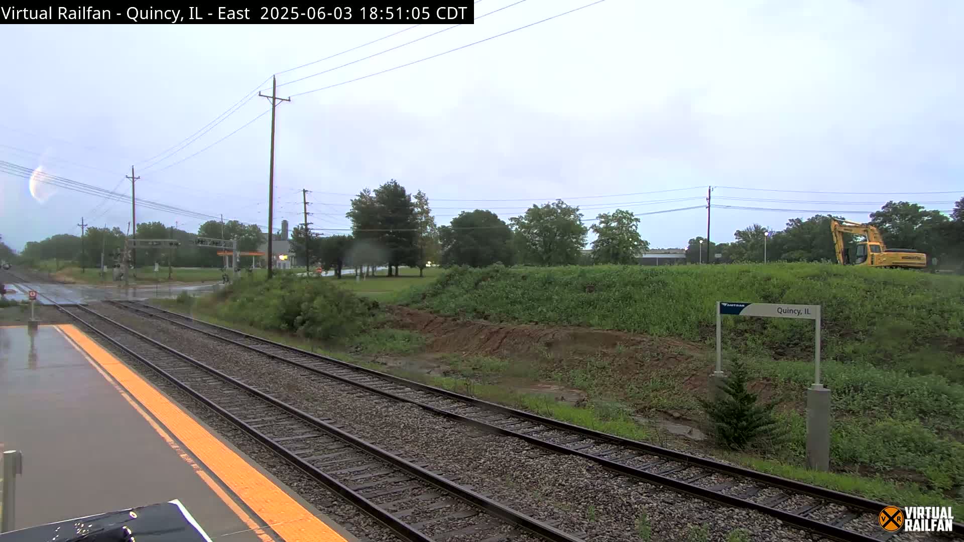 A wet train platform beside railroad tracks shows a partly cloudy sky and construction equipment in the background.