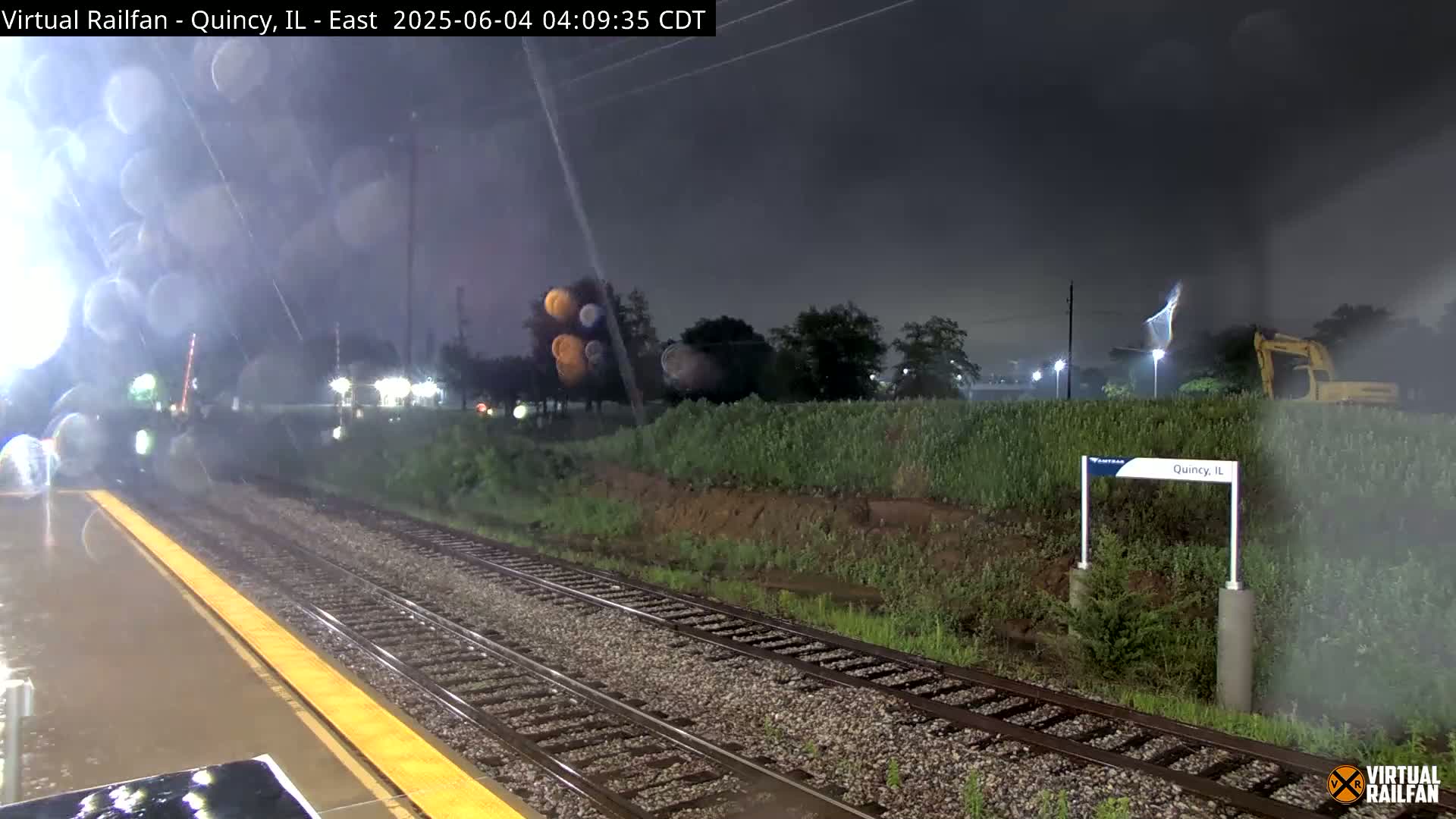 A nighttime view of rain-slicked train tracks and a platform, with a construction vehicle visible in the background under a dark, stormy sky.