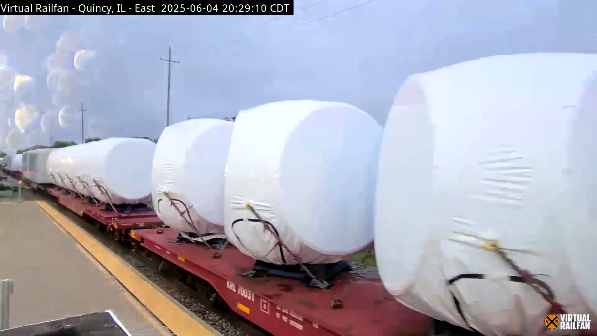 A long train carrying several large, white, cylindrical objects, wrapped in protective sheeting, moves along a track under an overcast sky.