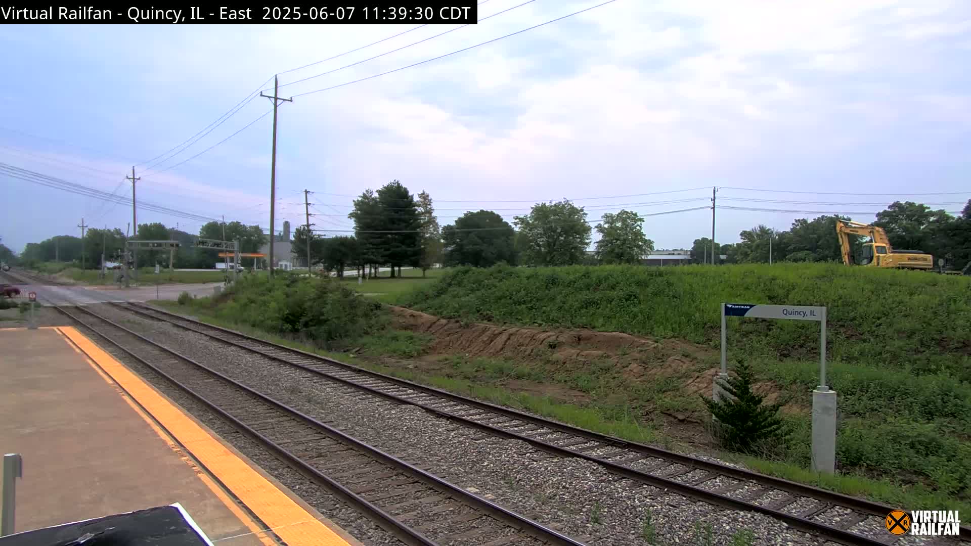 A train platform with two sets of railroad tracks extends into the distance under a partly cloudy sky, with a yellow excavator visible in the background beyond a grassy embankment.