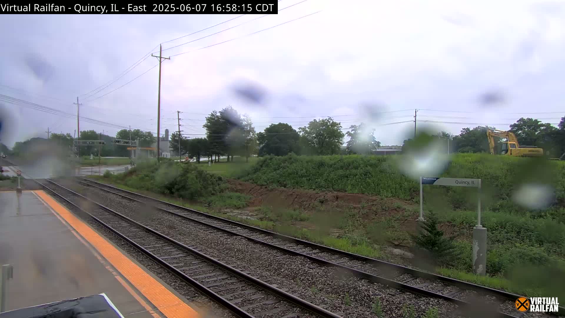 A rainy view of a train station platform and tracks, with a yellow excavator visible in the background.