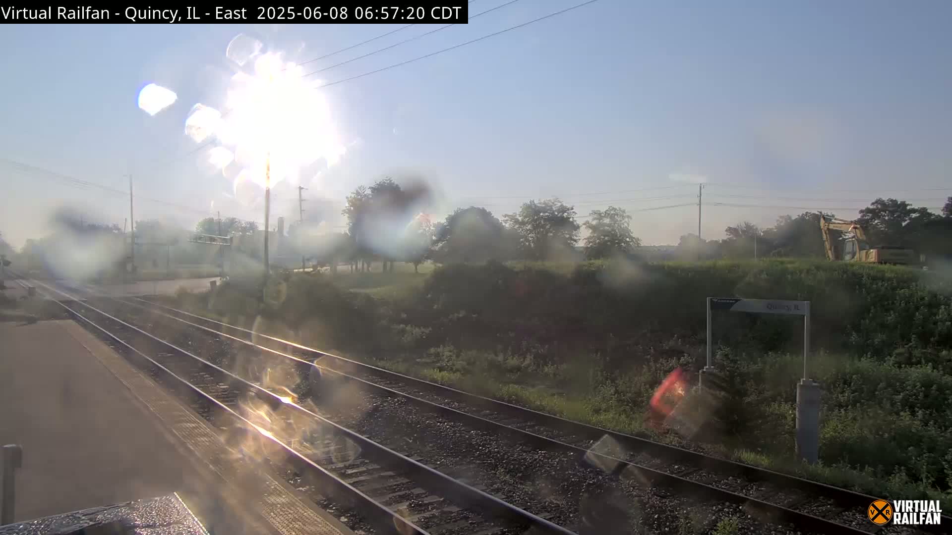 A view from a train shows railroad tracks and a sunny, slightly misty morning with vegetation alongside the tracks, including a piece of construction equipment in the distance.