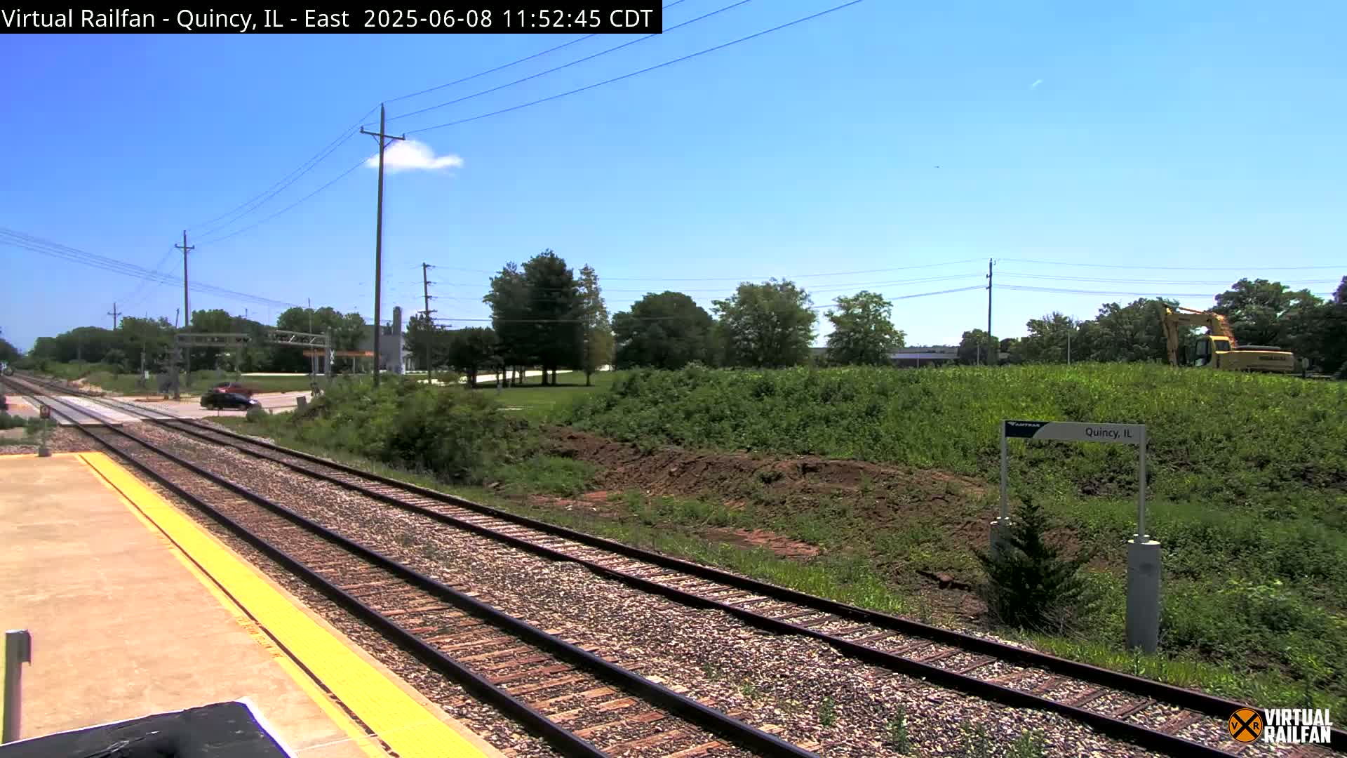 Under a mostly clear blue sky, a train track with a platform extends into the distance, passing a road intersection and a construction site with heavy machinery.