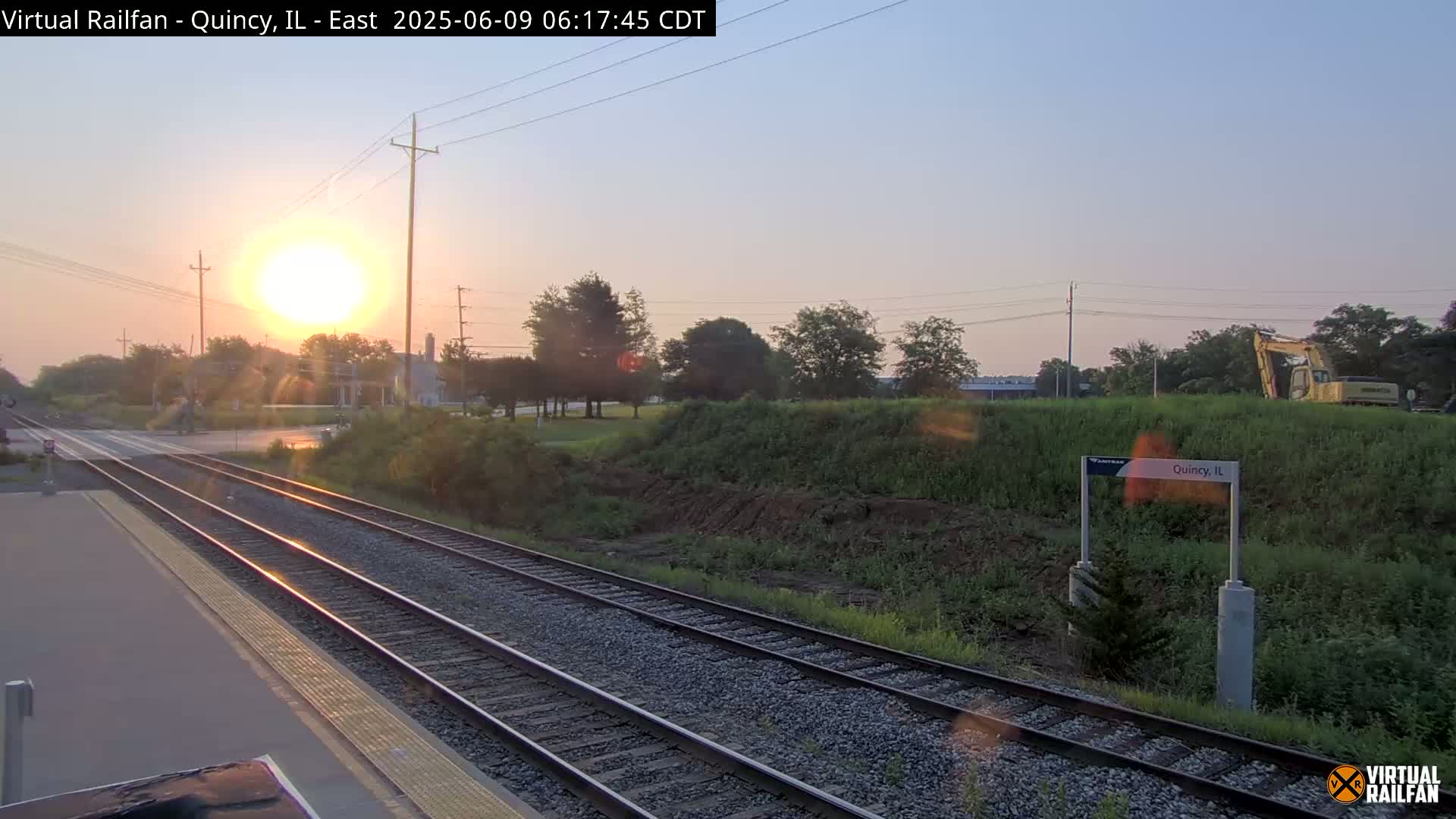 Sunrise illuminates a train station platform and tracks, with a construction vehicle visible beyond a grassy embankment.
