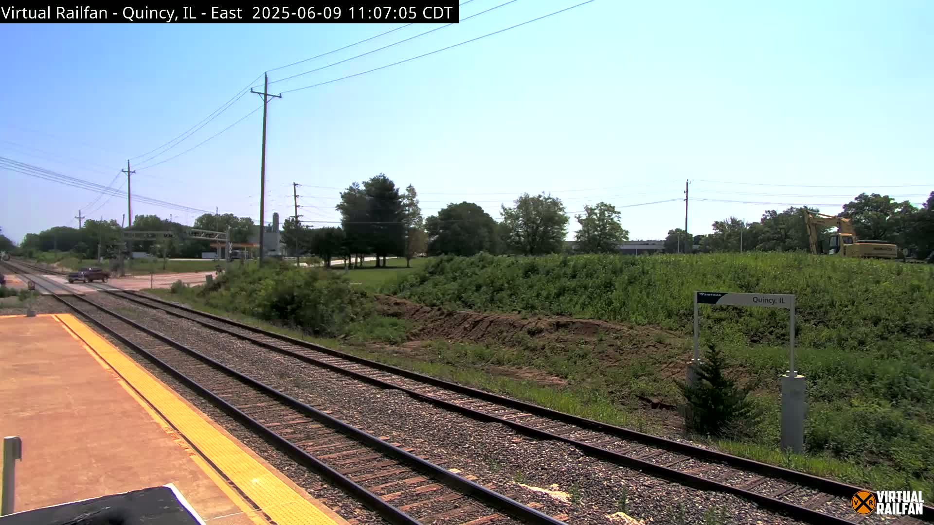 A train track with a platform stretches toward a road and some greenery under a clear, sunny sky.