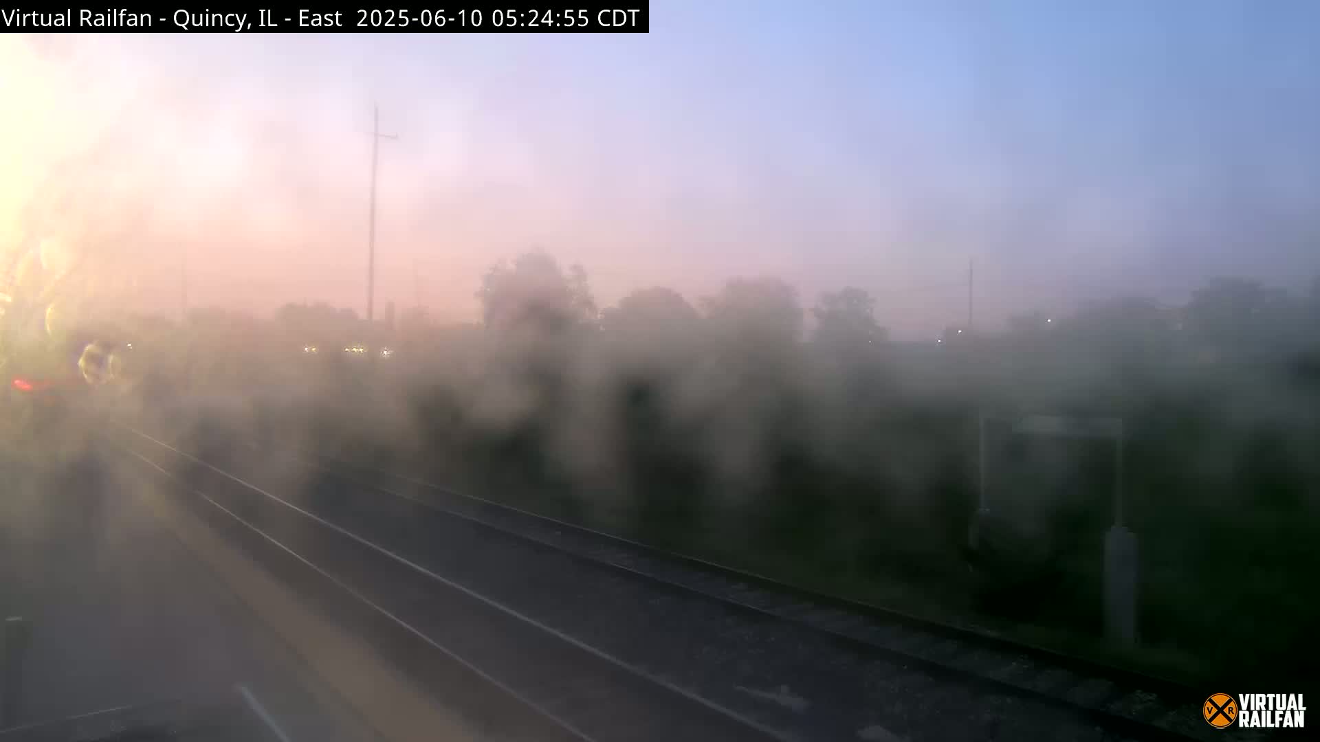 A foggy sunrise view from a train shows railroad tracks and vegetation beyond, obscured by moisture on the window.