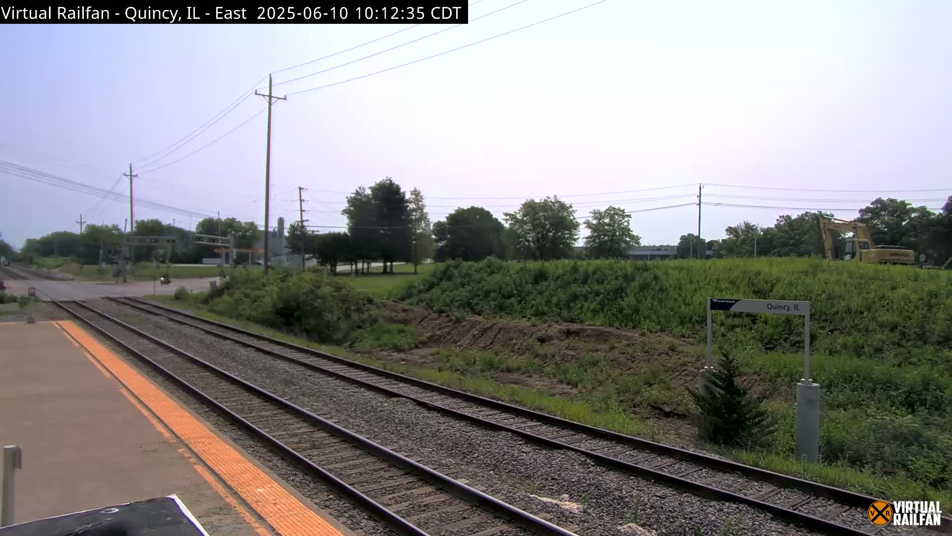 A train station platform with two sets of train tracks extends into the distance under a mostly clear sky, with some light cloud cover, beyond a grassy embankment where construction equipment is visible.