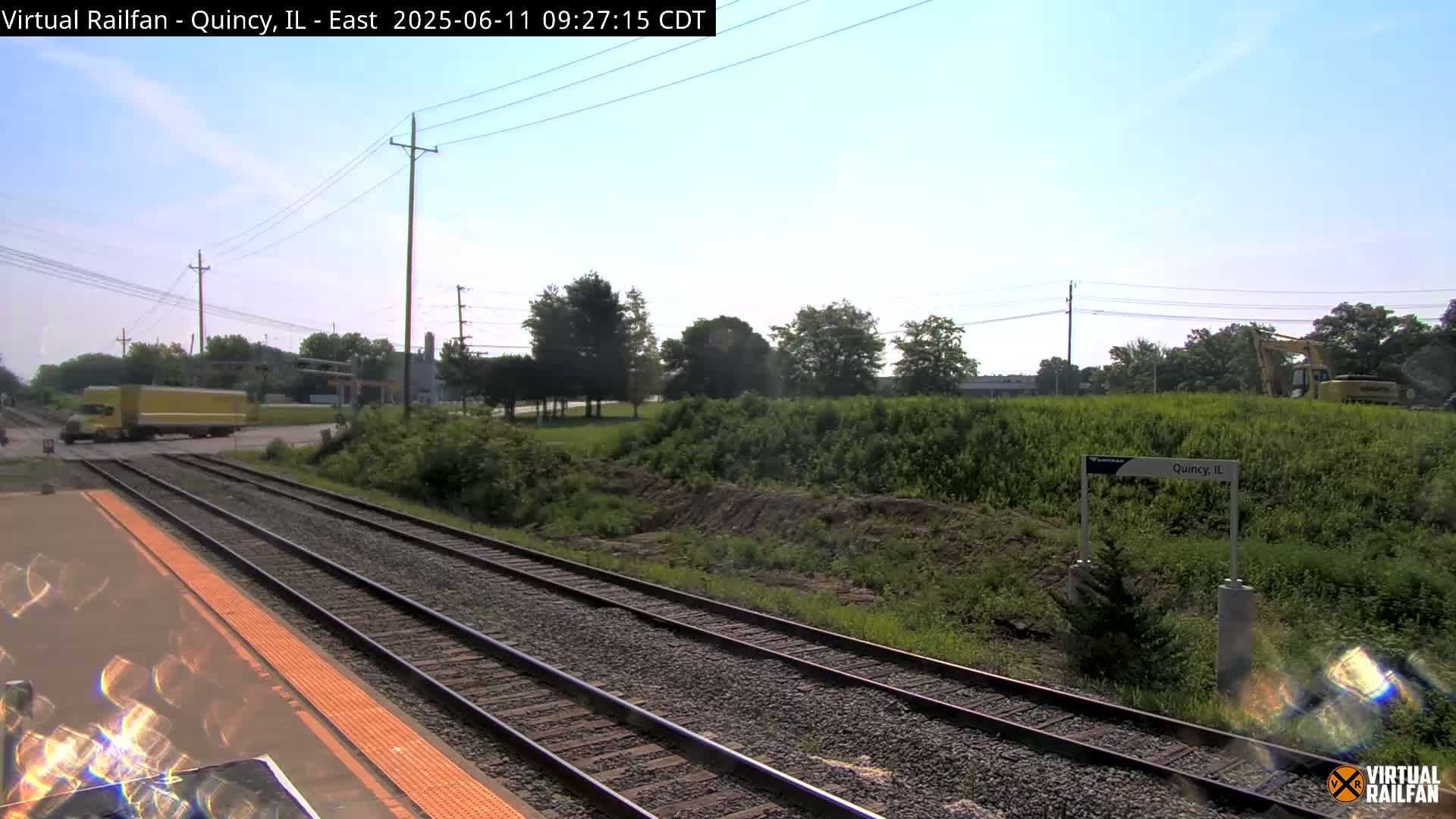 A yellow semi-truck crosses a railroad crossing near a train station on a sunny day.