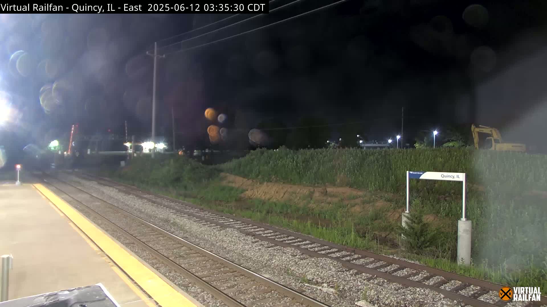 A nighttime view of a train platform with railroad tracks extending into the distance under a dark, hazy sky.