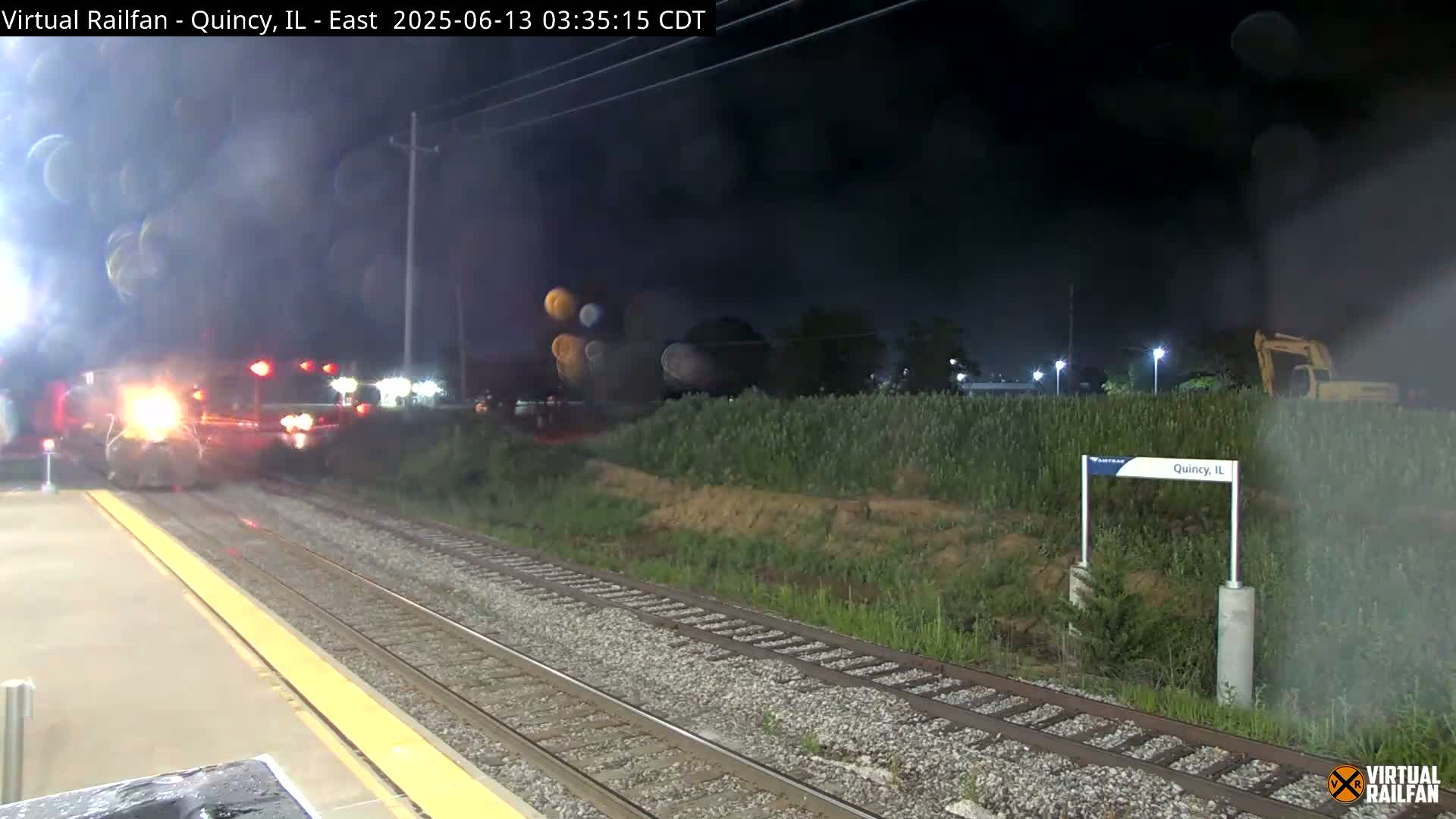 A train departs a nighttime station, passing a sign indicating Quincy, IL, under hazy conditions.
