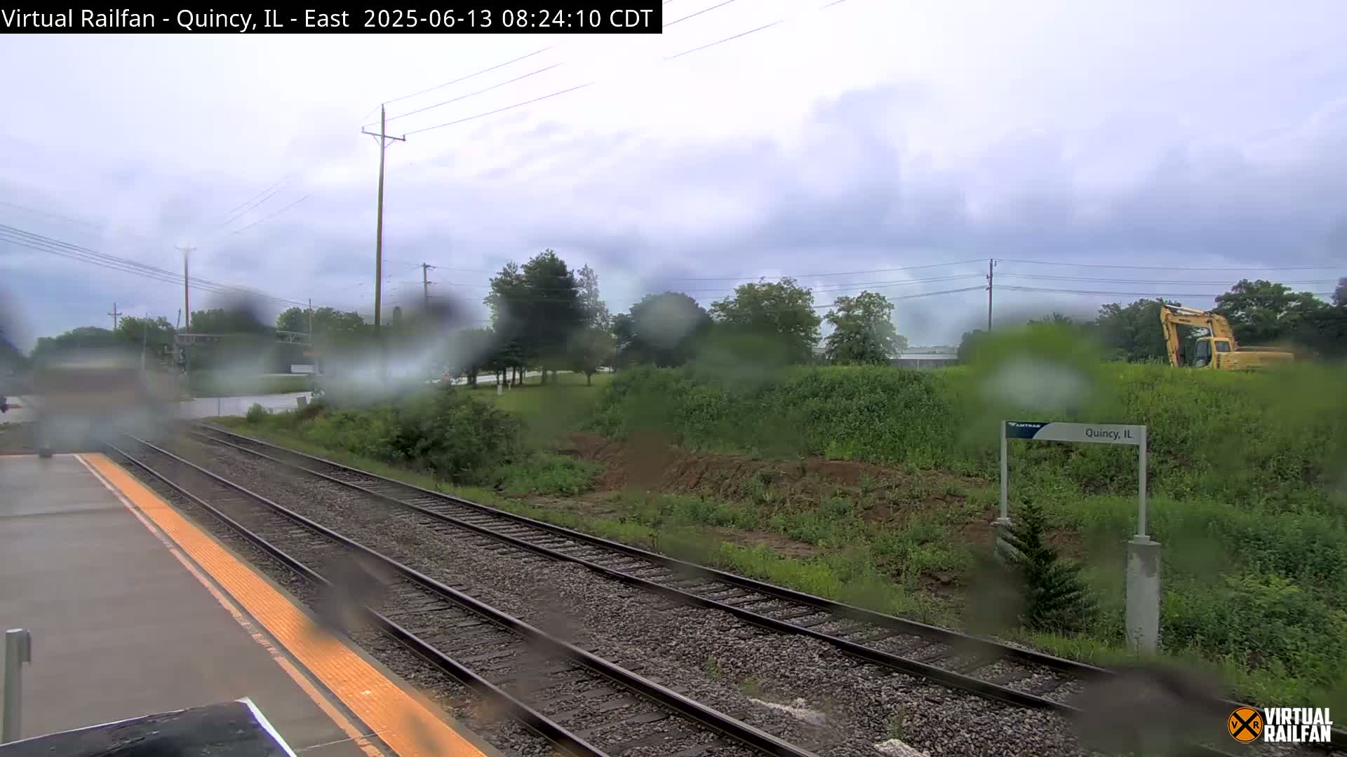 A rain-covered view of a train station platform and tracks, with a yellow excavator visible in the distance.
