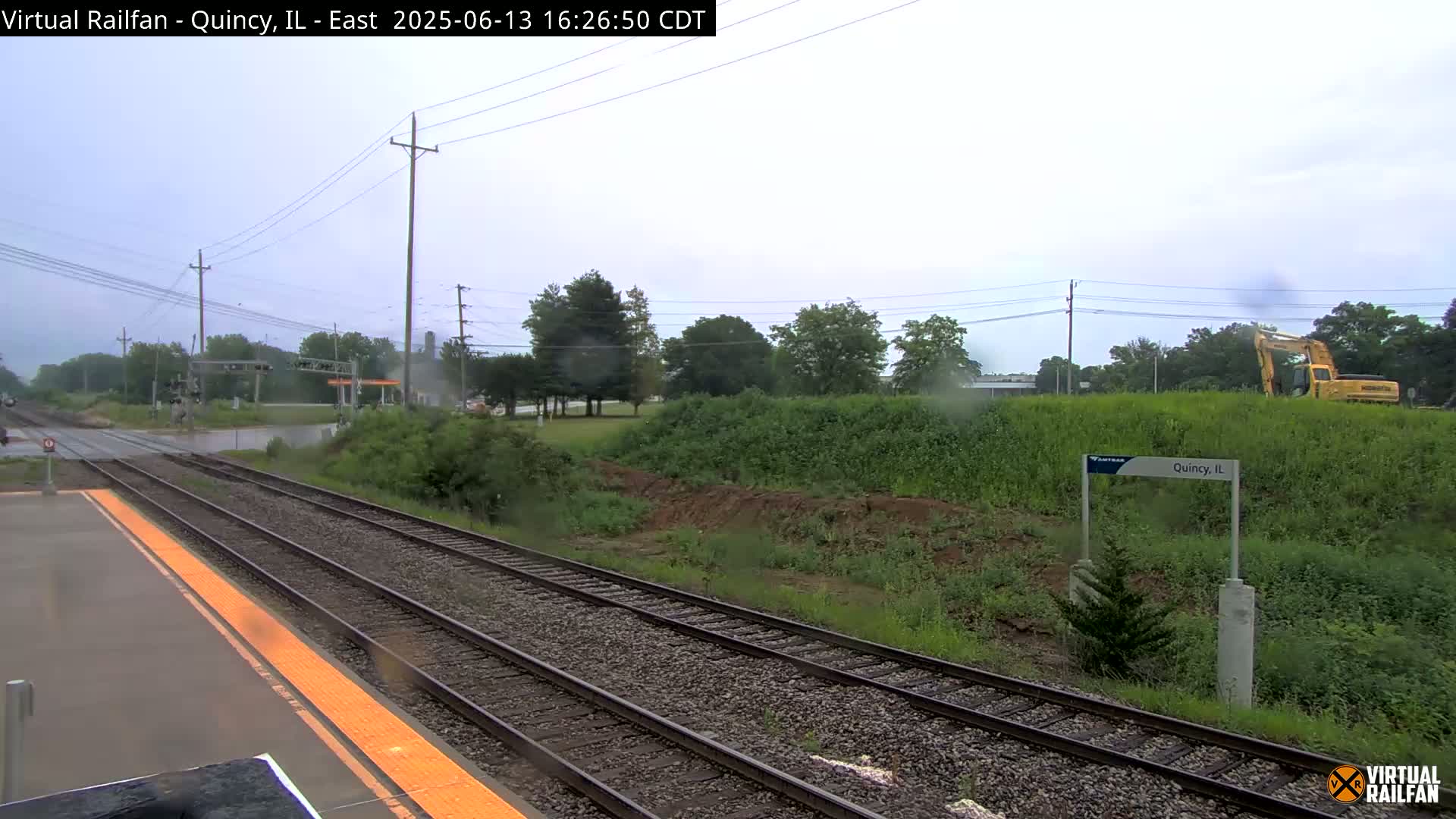 A view from a train shows a wet, overcast day at a train station with railroad tracks, greenery, and a construction vehicle in the distance.