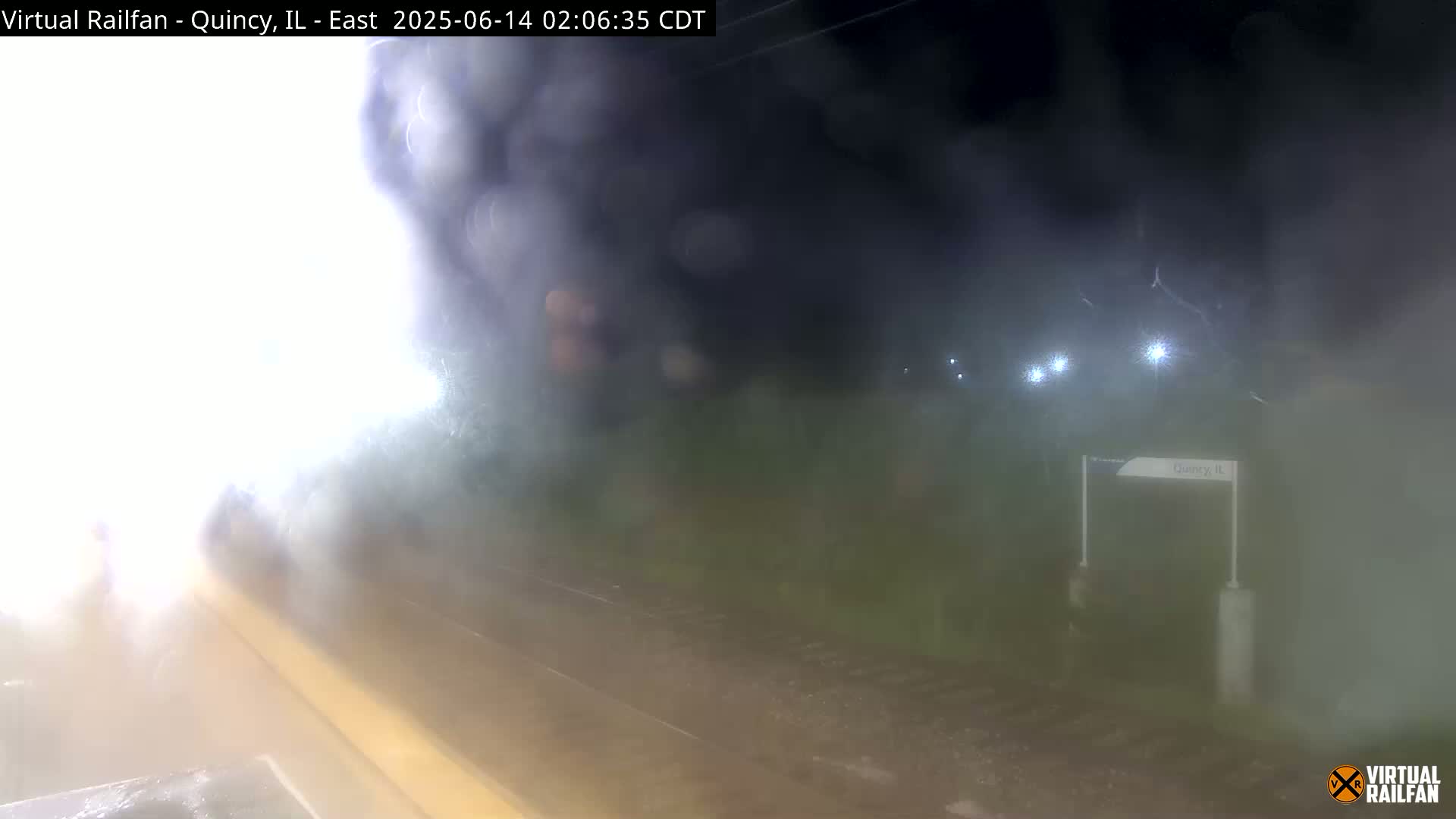 A nighttime view of a train station platform shrouded in fog, with lights illuminating the surroundings.