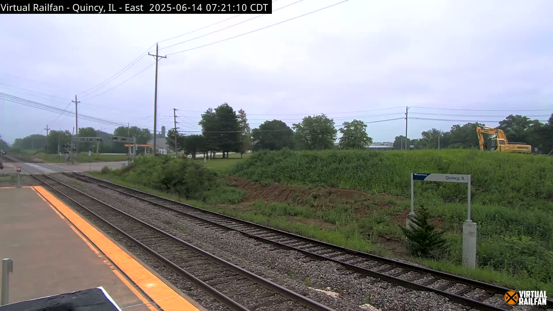 A train platform with multiple train tracks extends into the distance under an overcast sky, with some construction equipment visible beyond a grassy embankment.