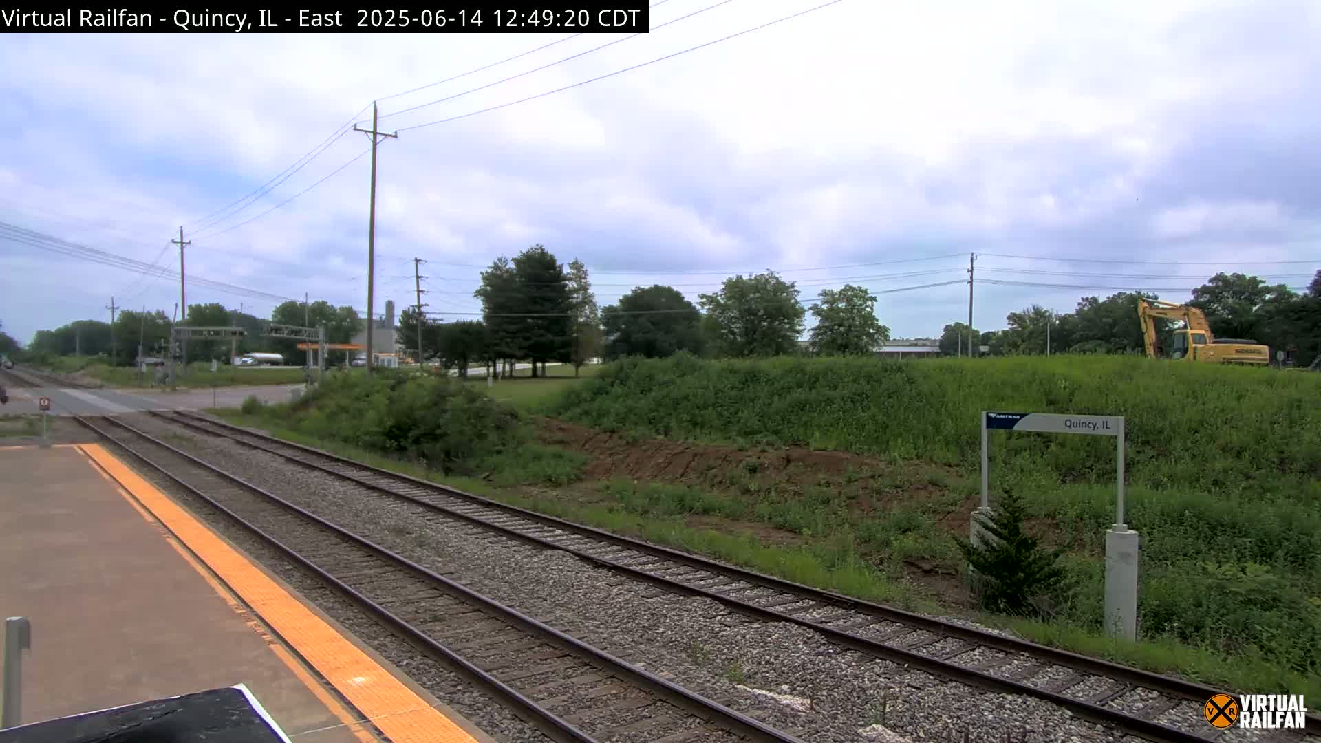 A train platform with tracks extending into the distance under a partly cloudy sky, next to a grassy embankment where some earthmoving is taking place.