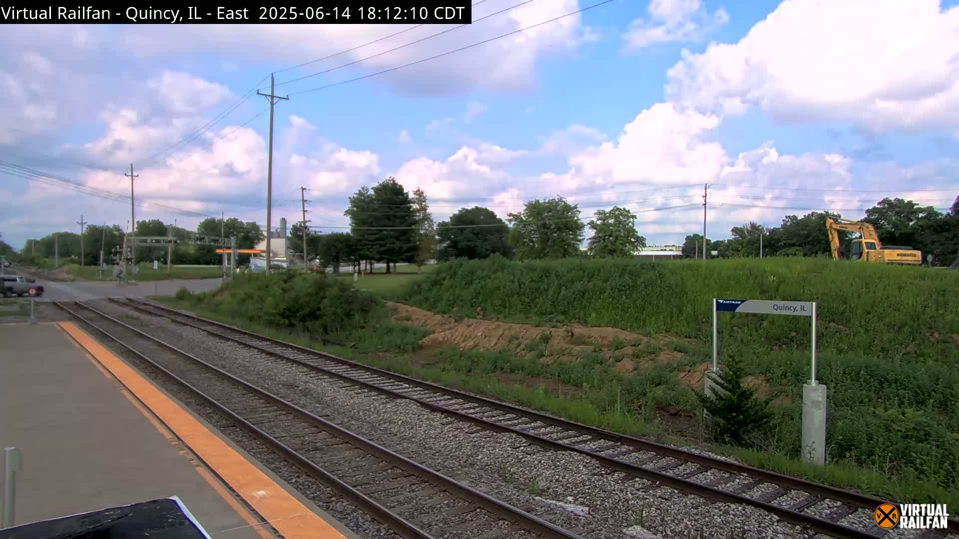 A train platform with tracks extending into the distance under a partly cloudy sky, next to a grassy embankment where some earthmoving is taking place.