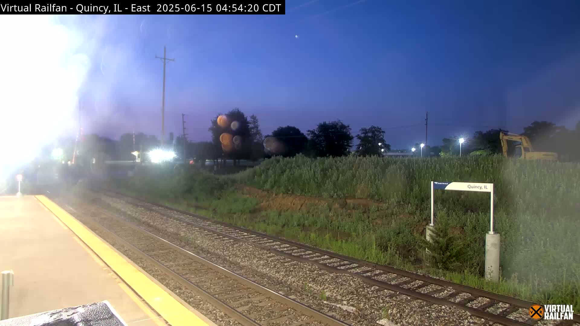 A train platform at dusk, with railroad tracks extending into the distance, is adjacent to a grassy area with a few trees and a piece of construction equipment.