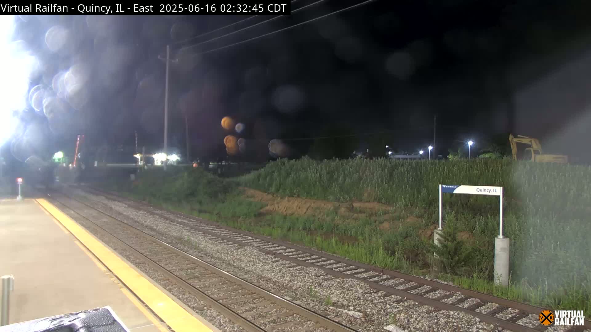 A nighttime view of train tracks at a station, with hazy or foggy conditions, and vegetation beside the tracks.