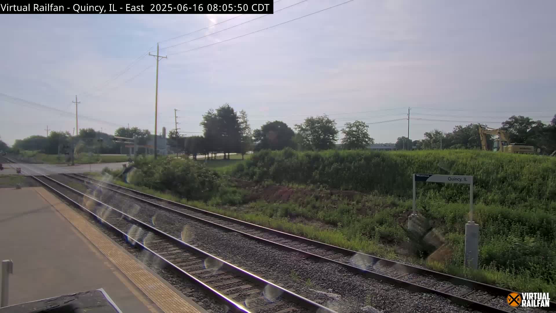 A train station platform with multiple train tracks stretches into the distance under a partly sunny sky.