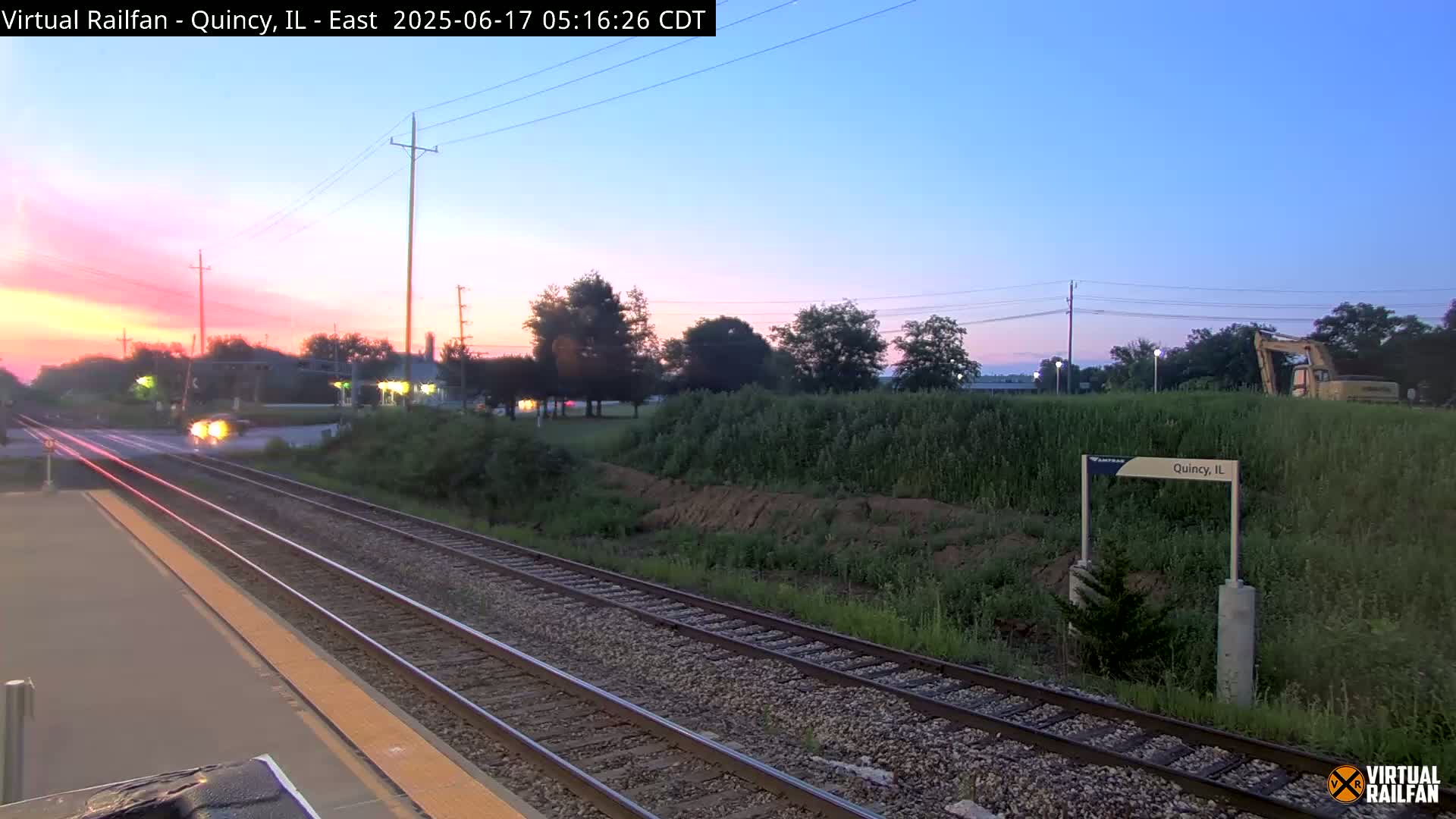 A sunrise illuminates a train platform with railroad tracks extending into the distance,  past some vegetation and construction equipment, under a clear sky.