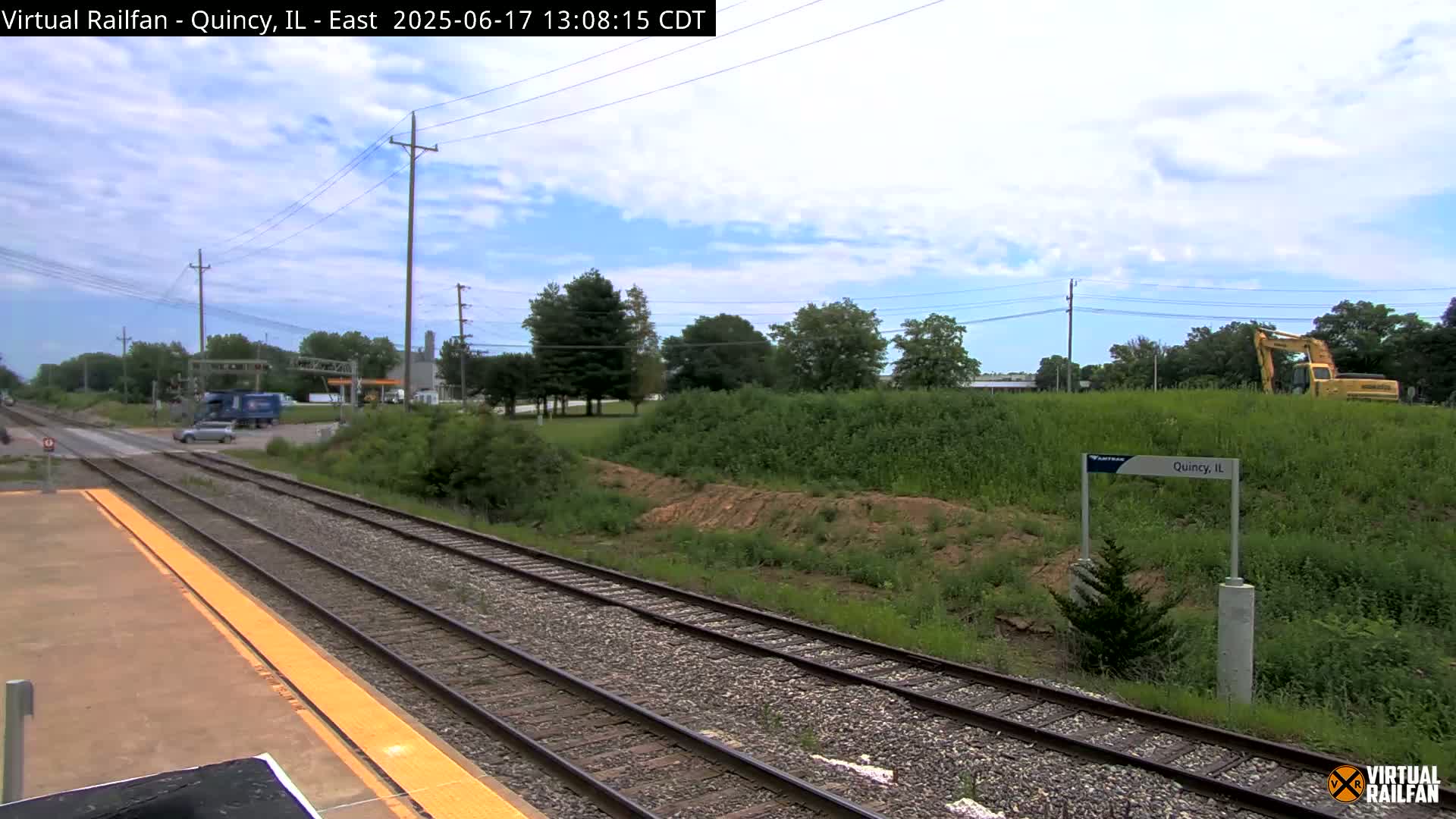 A partly cloudy sky overlooks a train platform with railroad tracks, a road crossing, some vegetation, and a yellow excavator in the distance.