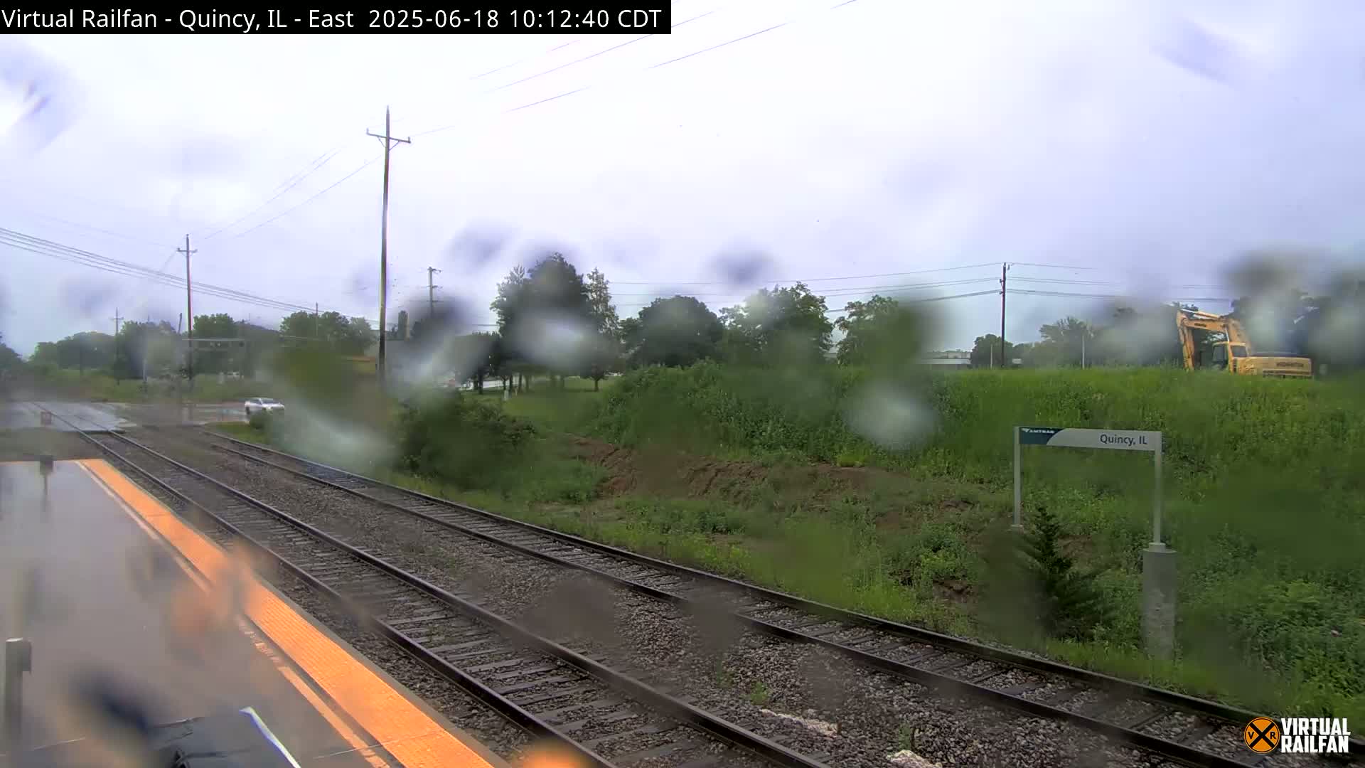 A rainy view of train tracks and a station platform, with a yellow excavator visible in the background.