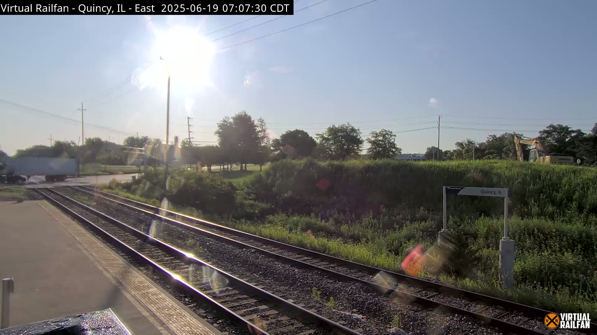 A sunny day at a train station shows multiple train tracks, a roadway crossing, some vegetation, and a piece of construction equipment in the distance.