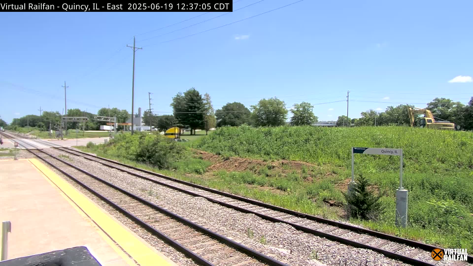 A sunny day reveals train tracks extending into the distance, alongside a grassy area with some earthworks and a small excavator, all under a clear blue sky.