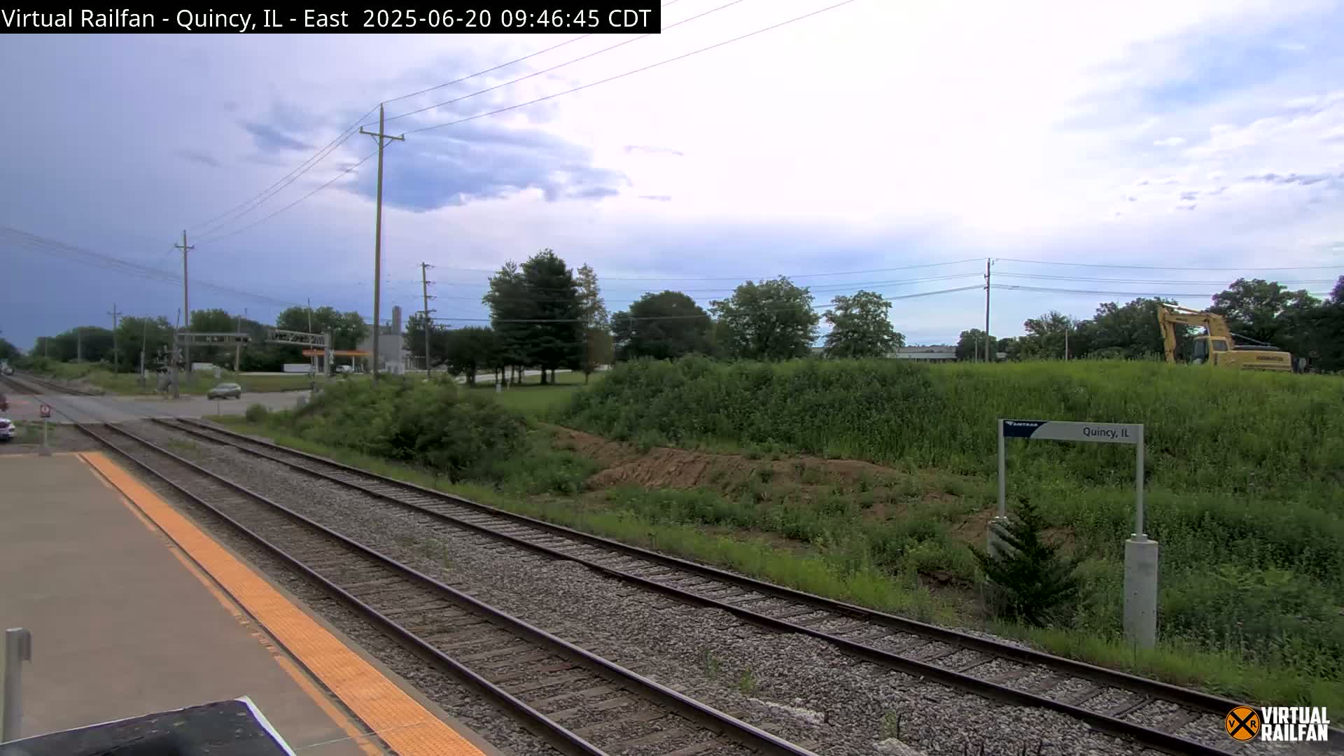 A view of train tracks and a nearby road, under a partly cloudy sky, with some construction equipment visible in the distance.