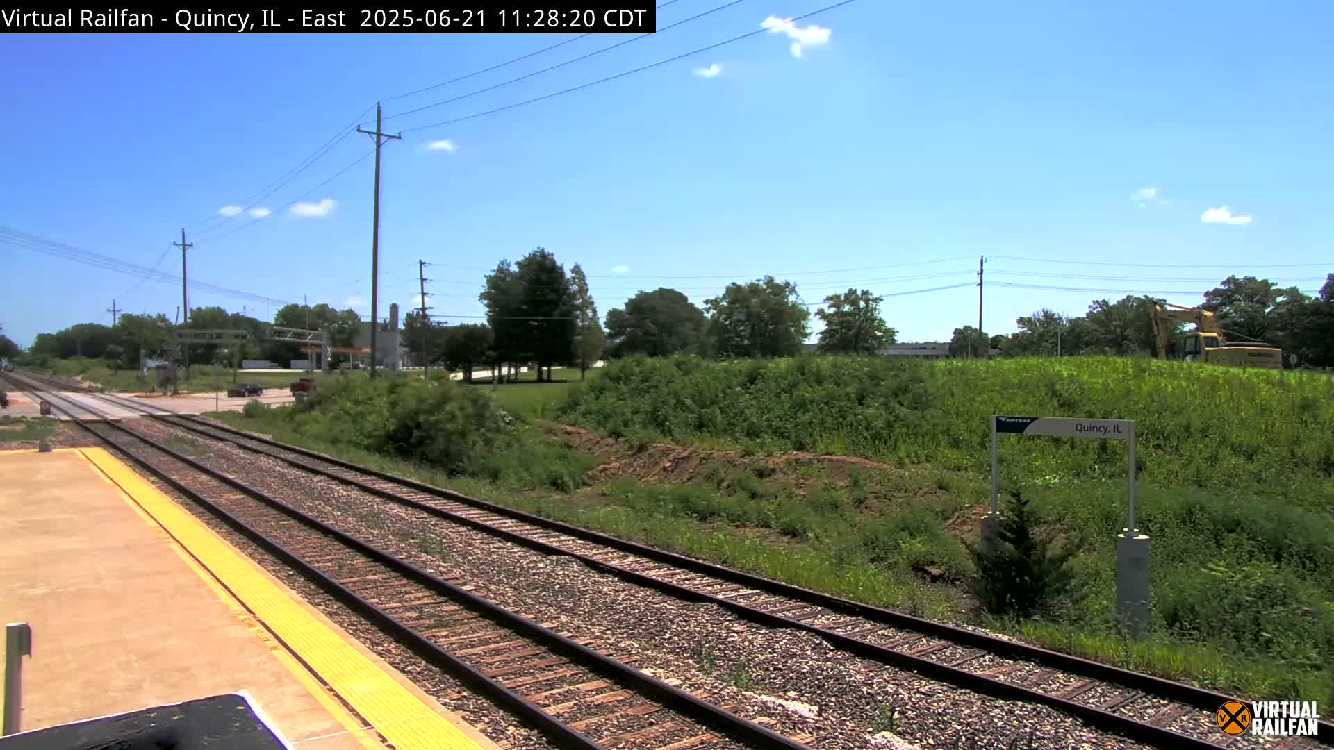 A sunny day shows a view of train tracks extending into the distance, bordered by green vegetation and a few utility poles, with a piece of construction equipment visible in the background.
