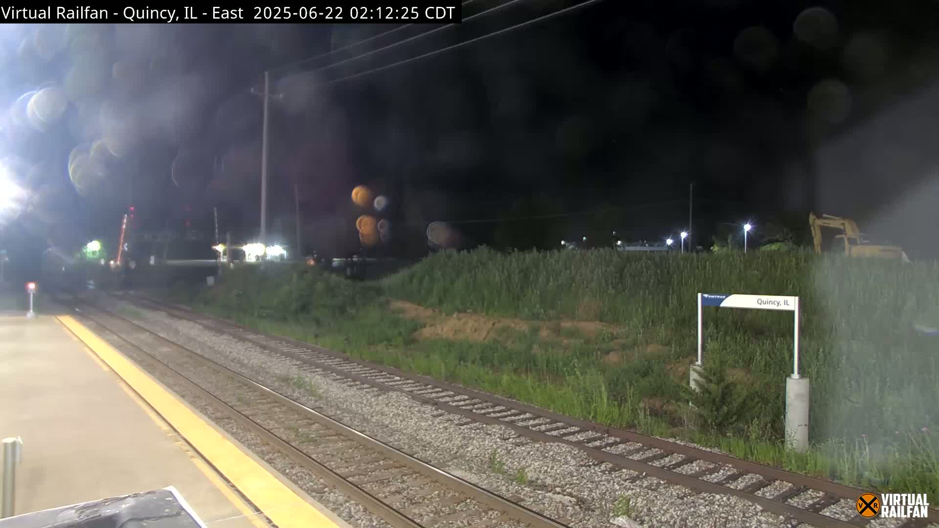 A nighttime view of train tracks at a station, with a hazy atmosphere and overgrown vegetation beside the tracks.