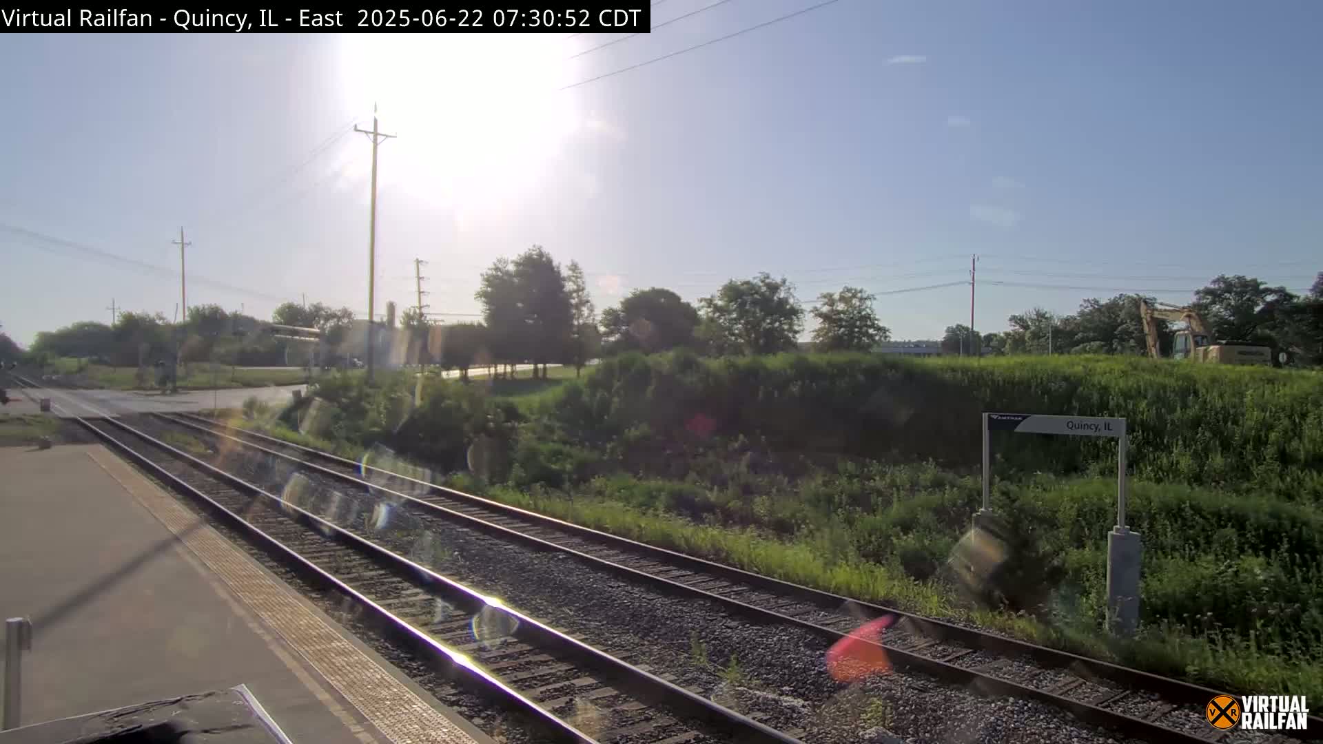 A sunny day view of train tracks and a station platform, with green vegetation and a construction vehicle visible in the distance.