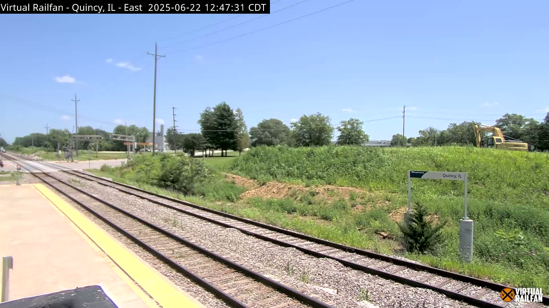 A sunny day view of train tracks extending into the distance, alongside a grassy area with some earthworks and a piece of construction equipment.