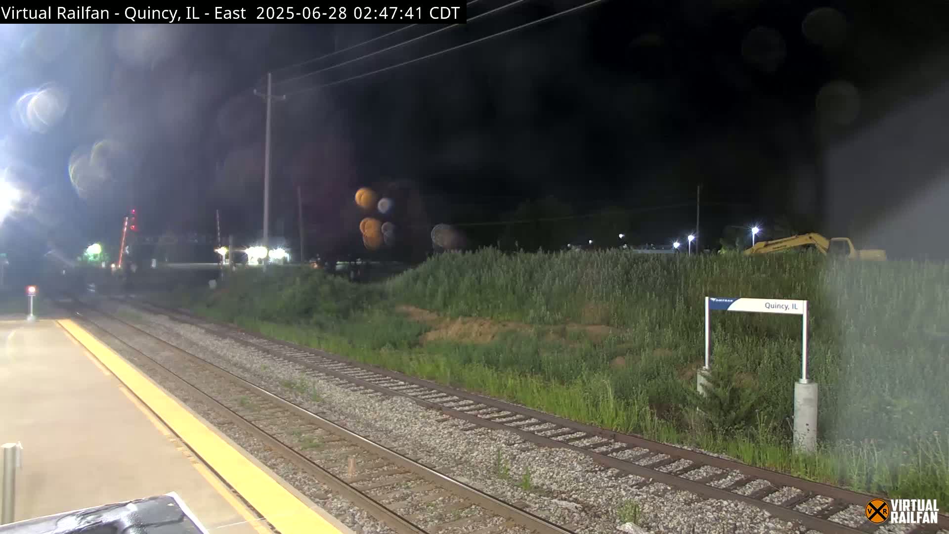 A nighttime view of a train station platform with railroad tracks extending into a weedy area, under a hazy sky.