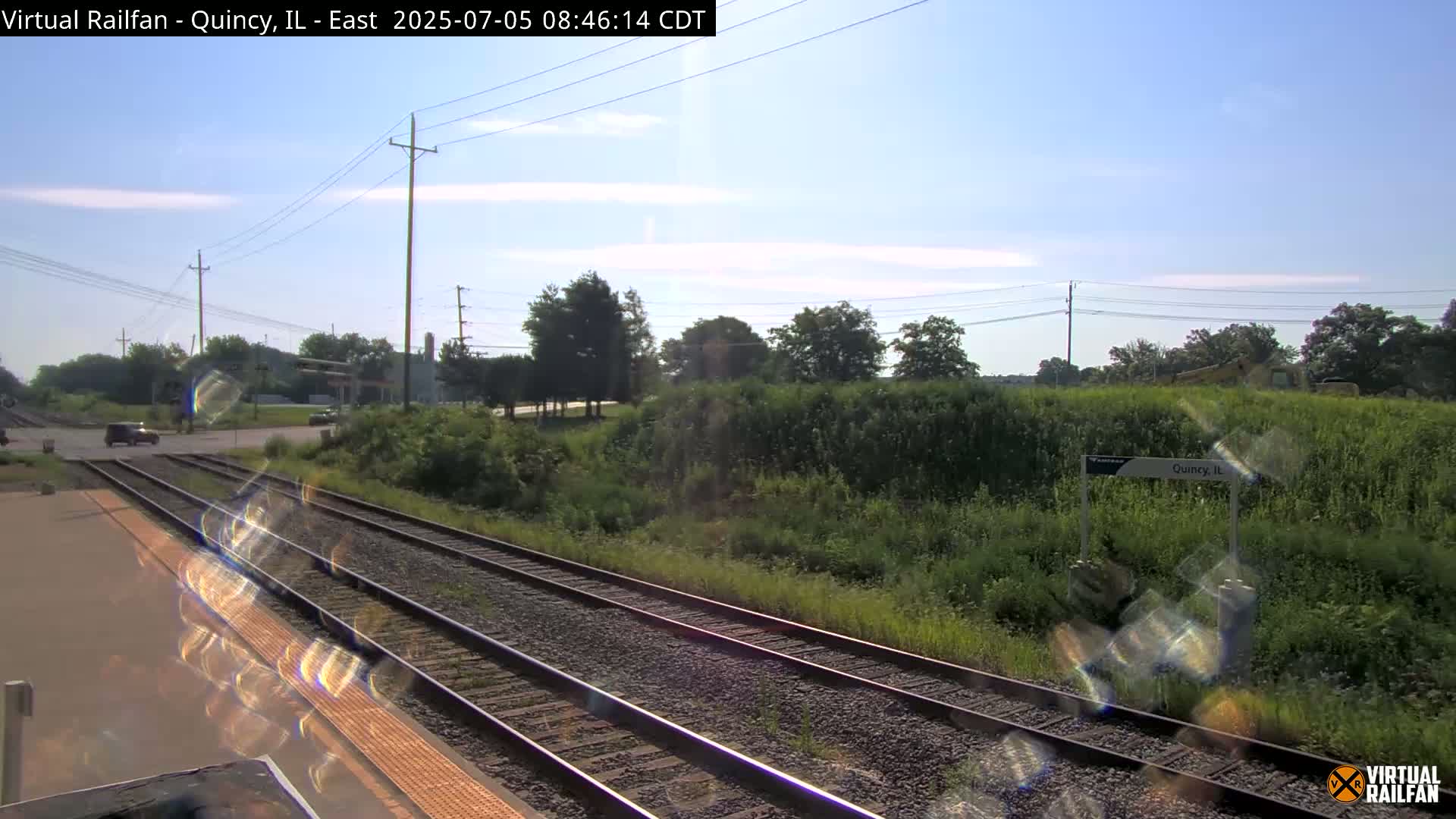 A sunny day shows a train station platform next to railroad tracks, with green vegetation and a road visible in the background.