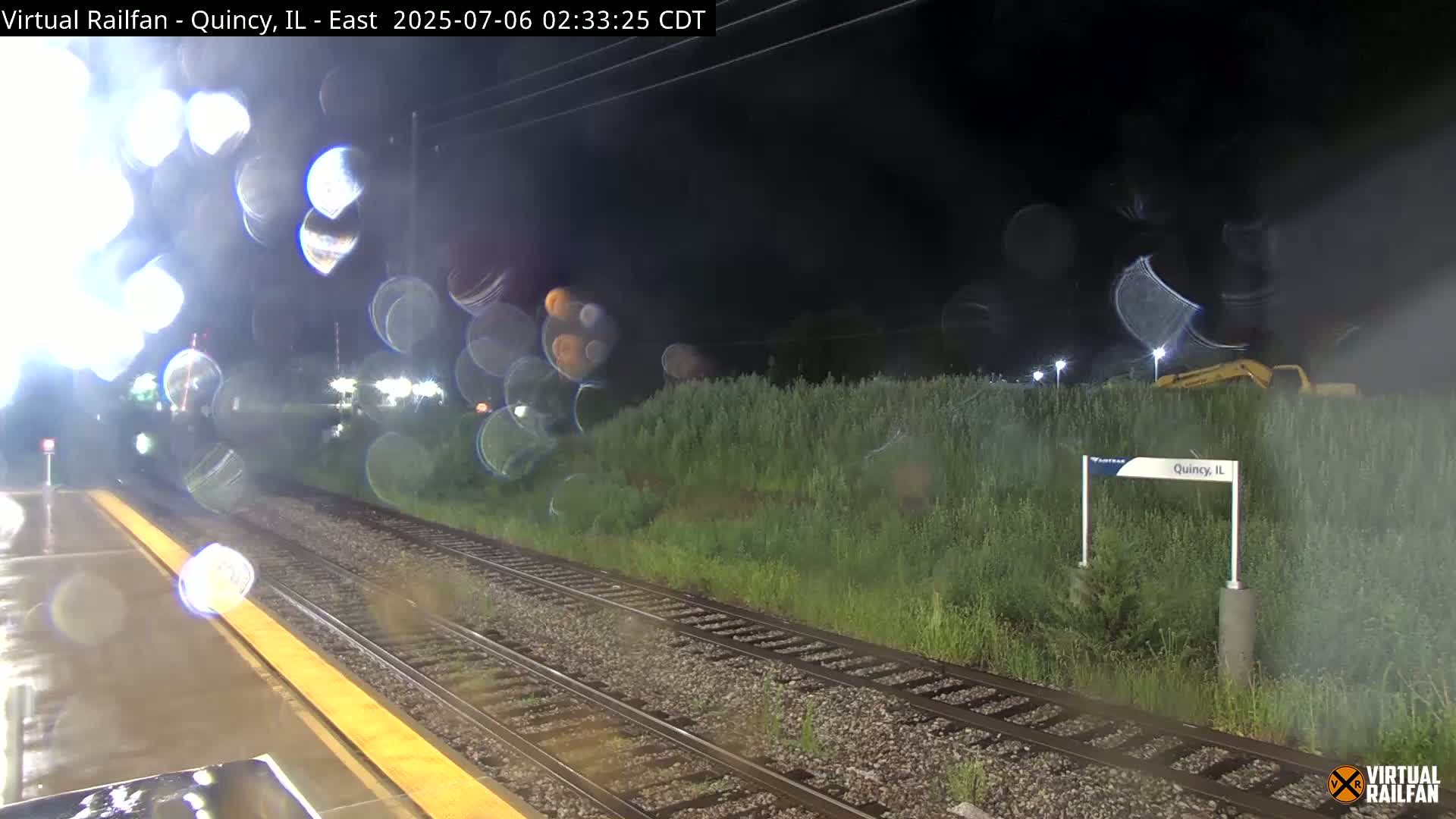 A nighttime view of a rain-slicked train platform and tracks, with overgrown vegetation beside them, and a piece of heavy machinery visible in the distance.