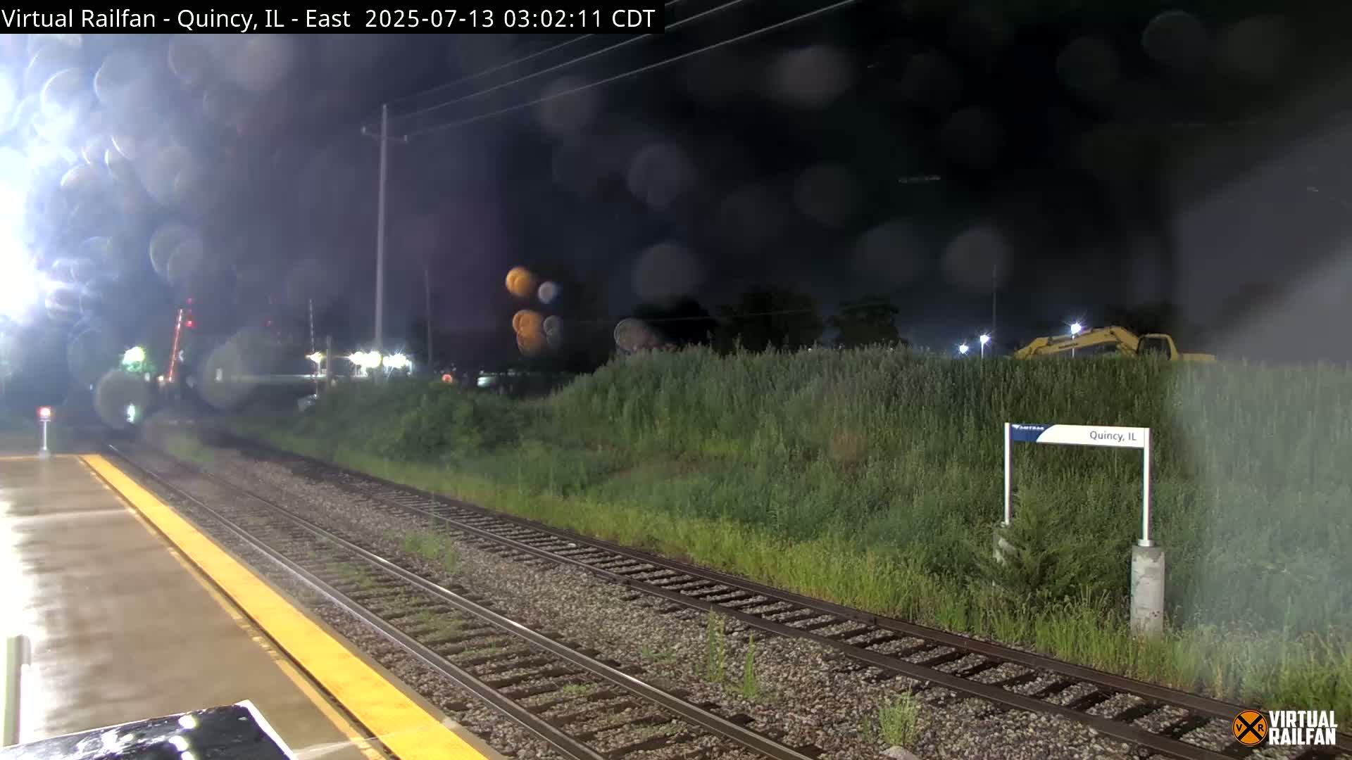 A nighttime view of a rain-slicked train platform and tracks, with overgrown vegetation and construction equipment visible beyond.