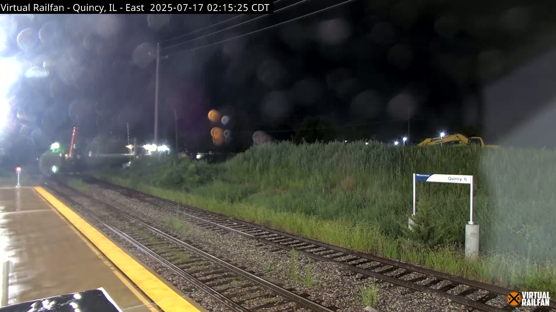 A nighttime view of rain-slicked train tracks and a platform, with overgrown vegetation and a piece of yellow construction equipment visible beyond.