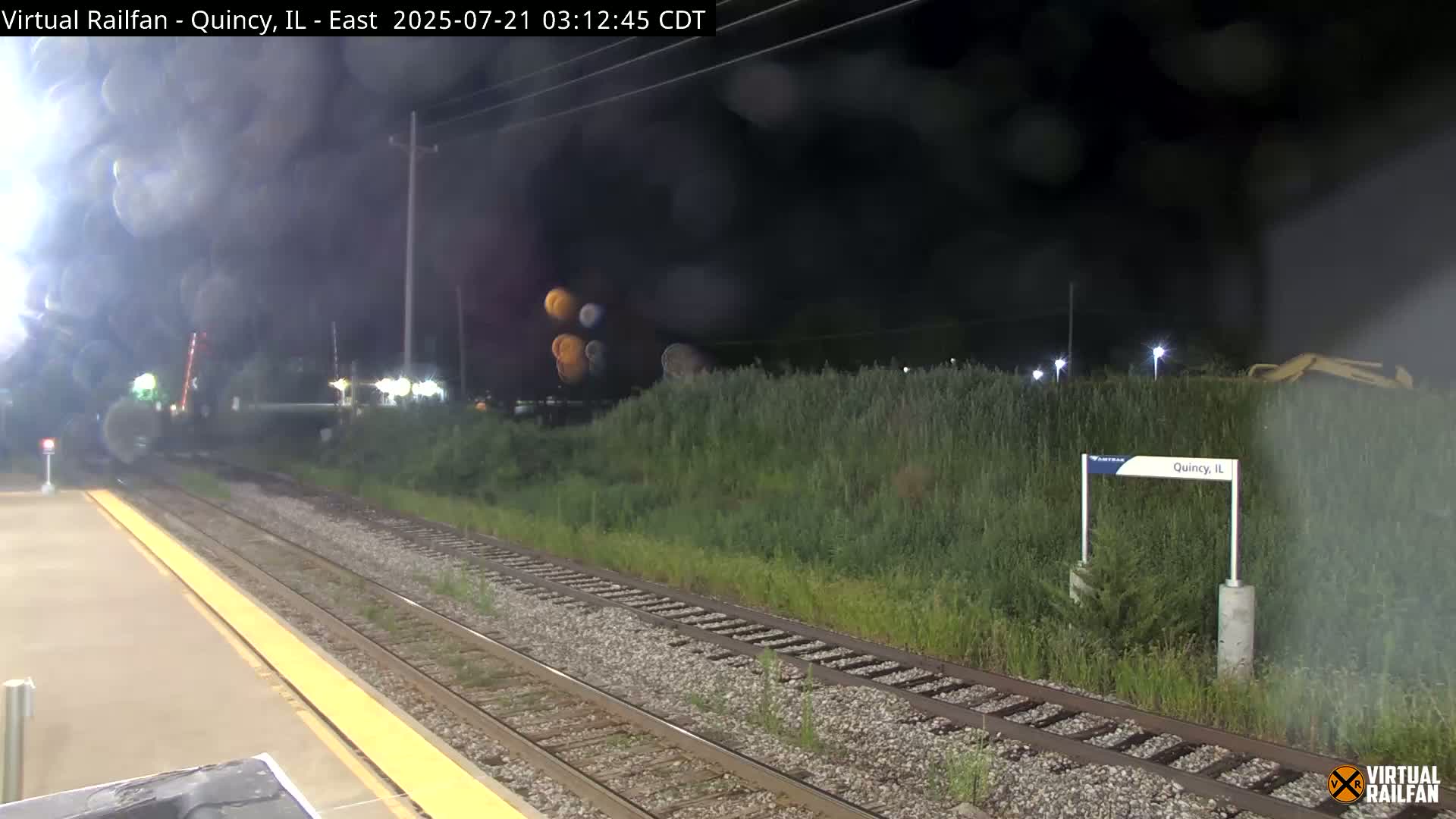 A nighttime view of a train station platform with railroad tracks, overgrown vegetation, and a train in the distance under a dark, cloudy sky.