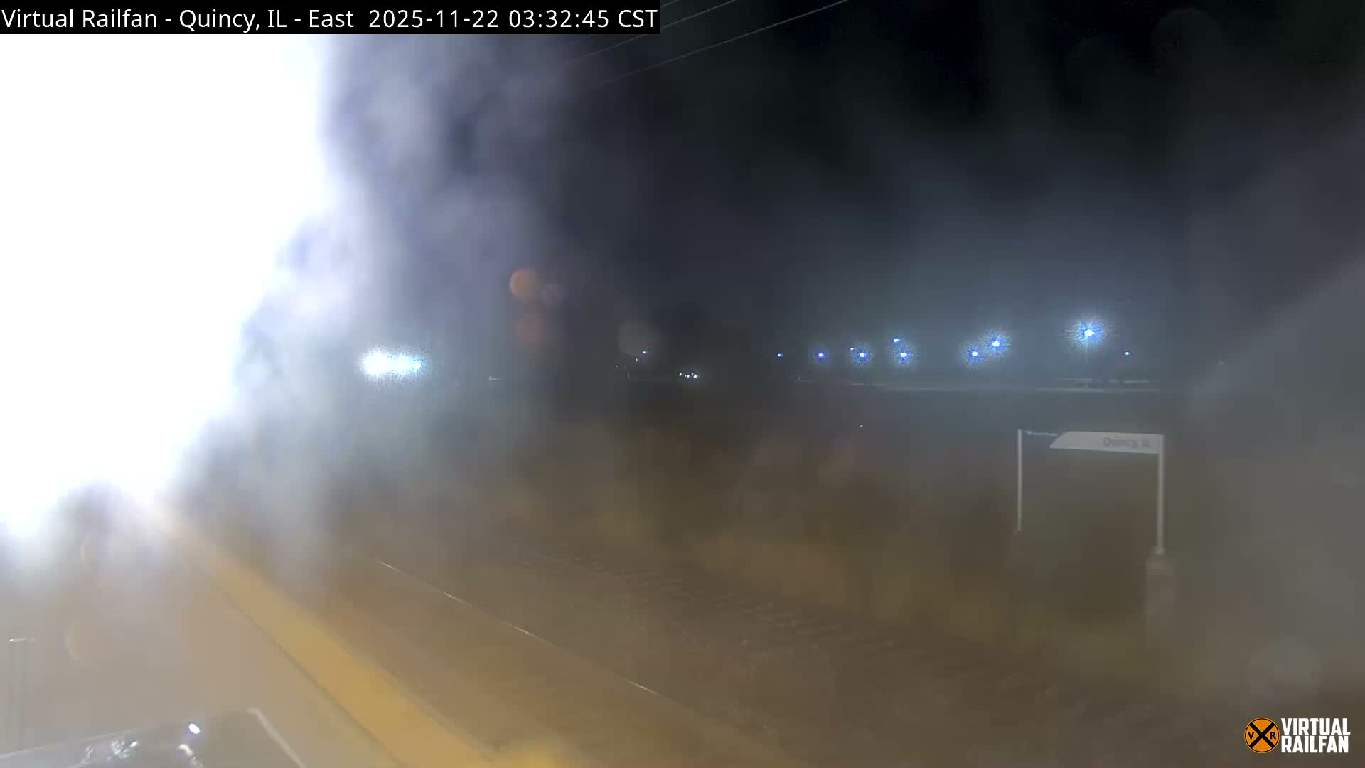 A nighttime outdoor scene at a railroad appears heavily obscured by dense fog or smoke, with tracks and a platform visible in the foreground, and distant streetlights glowing dimly through the pervasive haze.