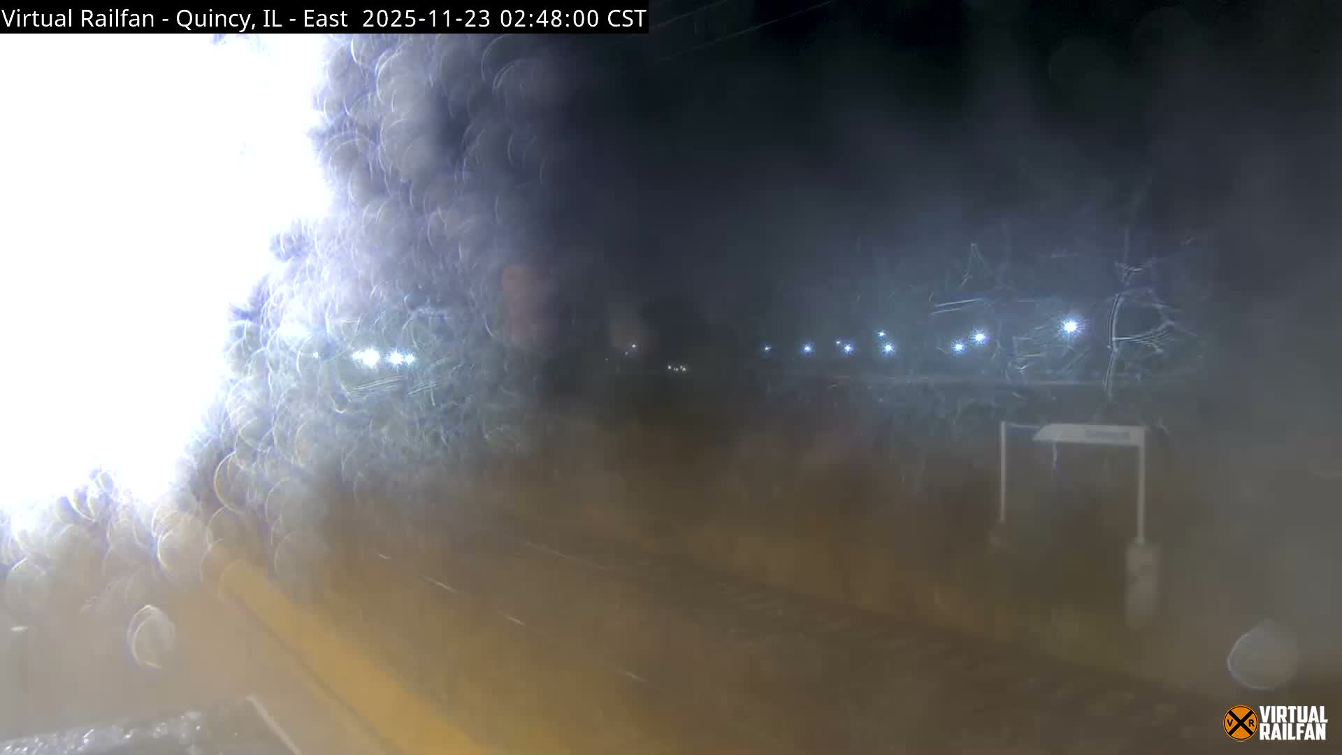 The image shows a dark, blurry outdoor scene at night, likely a railway, heavily obscured by rain or snow, with distant lights piercing through the precipitation.