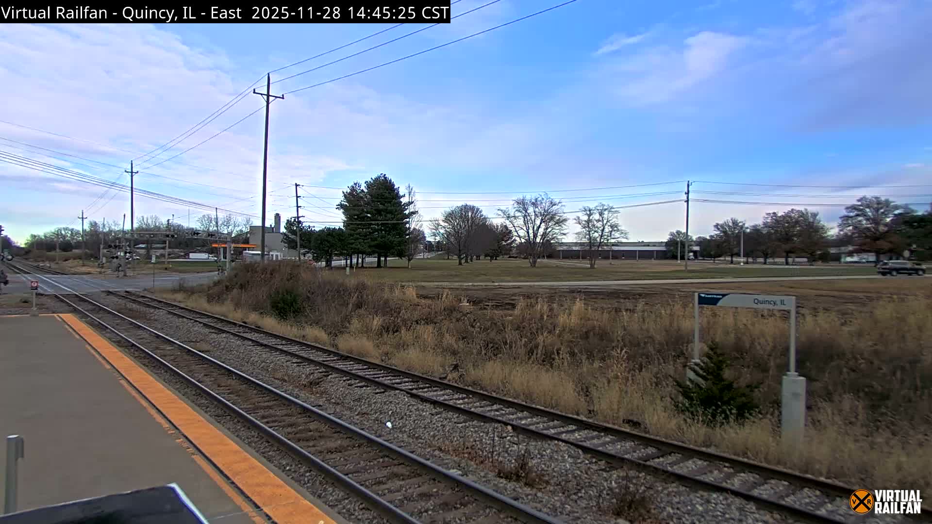 An outdoor scene captures multiple train tracks running alongside a concrete platform with a yellow safety strip, leading into a crossing and surrounded by dry, brown grasses, a mix of deciduous and evergreen trees, and distant industrial buildings under a bright blue sky with scattered white clouds.