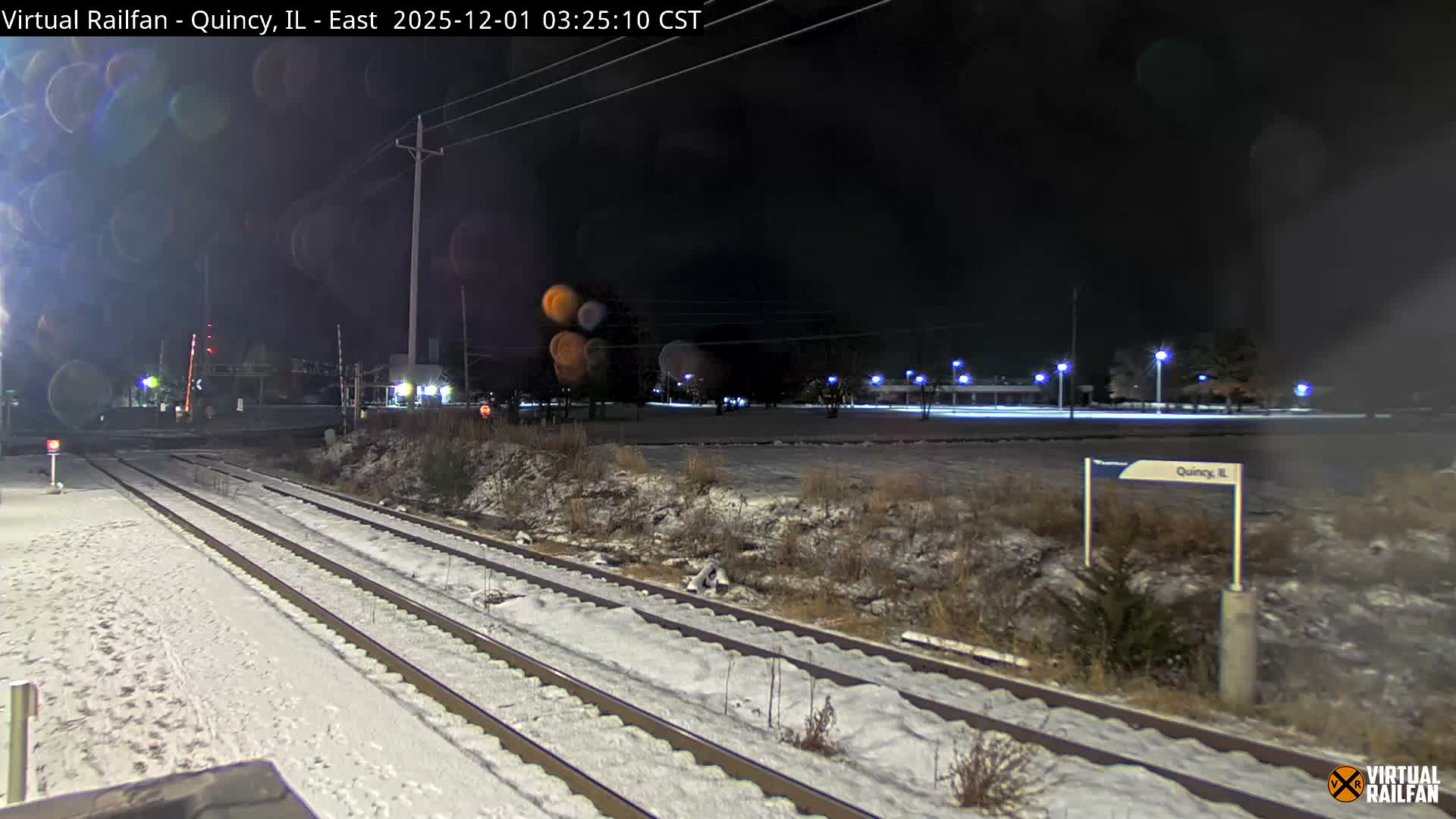 A snow-covered railway line with multiple tracks and an illuminated railroad crossing is seen at night, under a dark sky with artificial lights, indicating clear but snowy and cold weather.