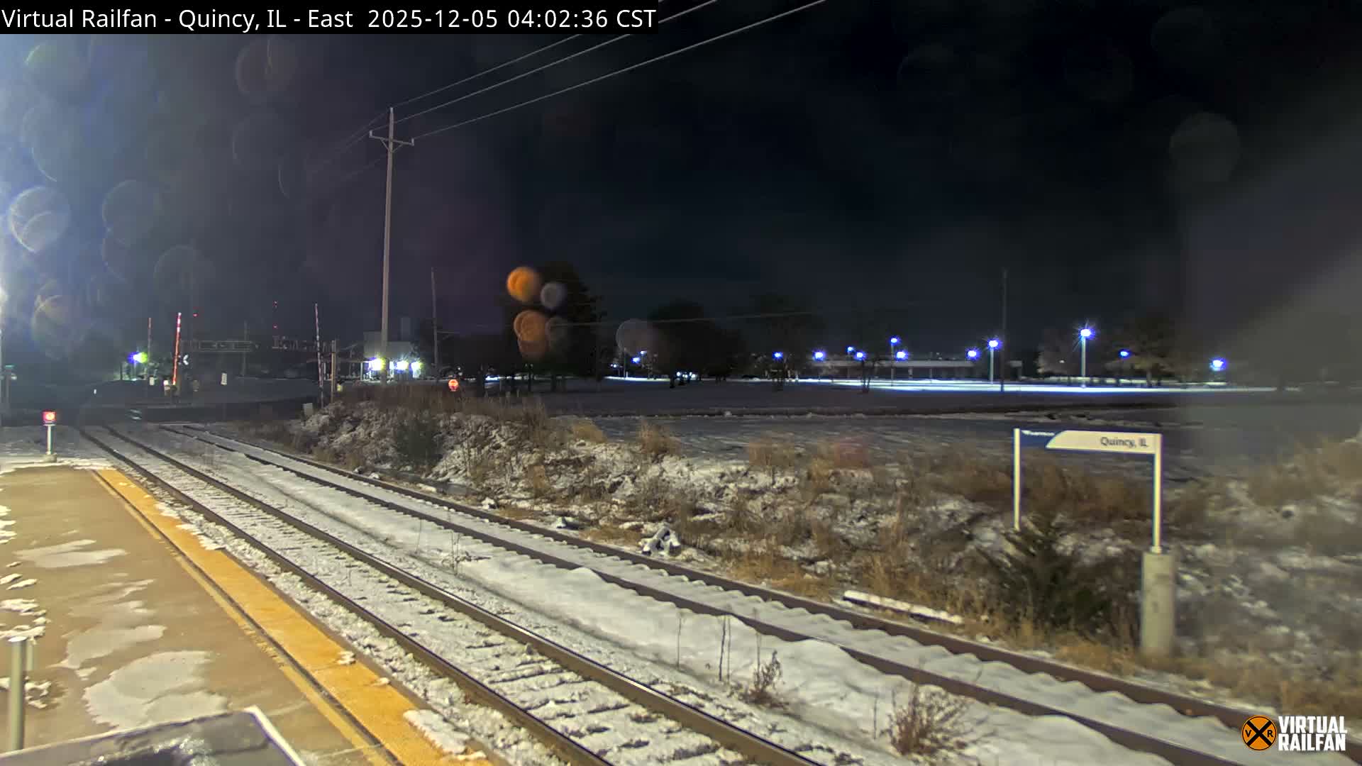 A nighttime view of a snowy railway scene reveals two tracks and a platform alongside a railroad crossing, with distant streetlights illuminating the cold winter landscape.