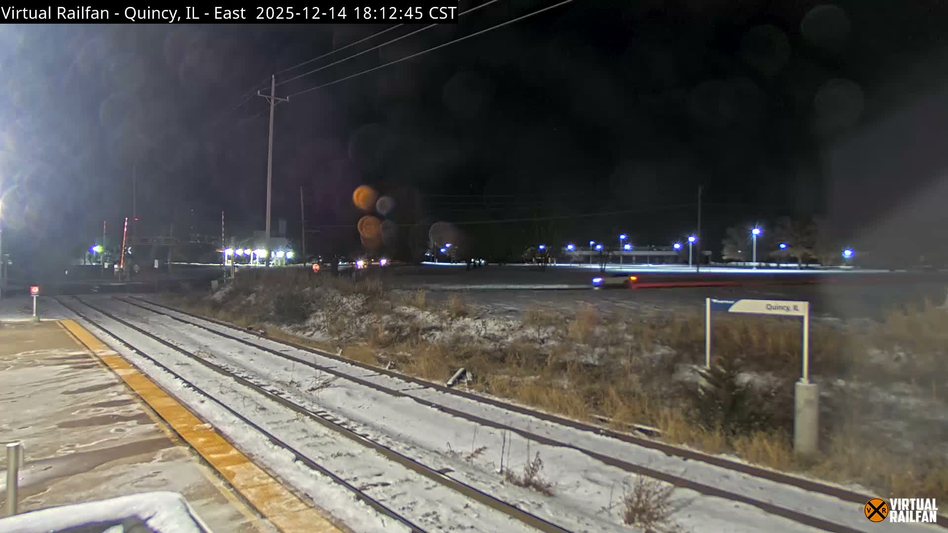 A nighttime view of a snowy railway scene reveals two tracks and a platform alongside a railroad crossing, with distant streetlights illuminating the cold winter landscape.