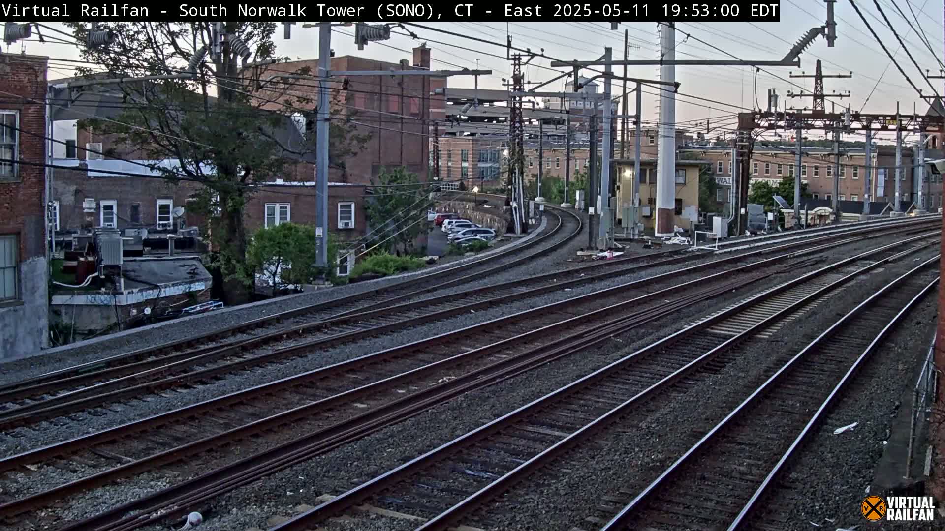 Multiple sets of train tracks curve gently to the left, passing by buildings and parked cars under a twilight sky.