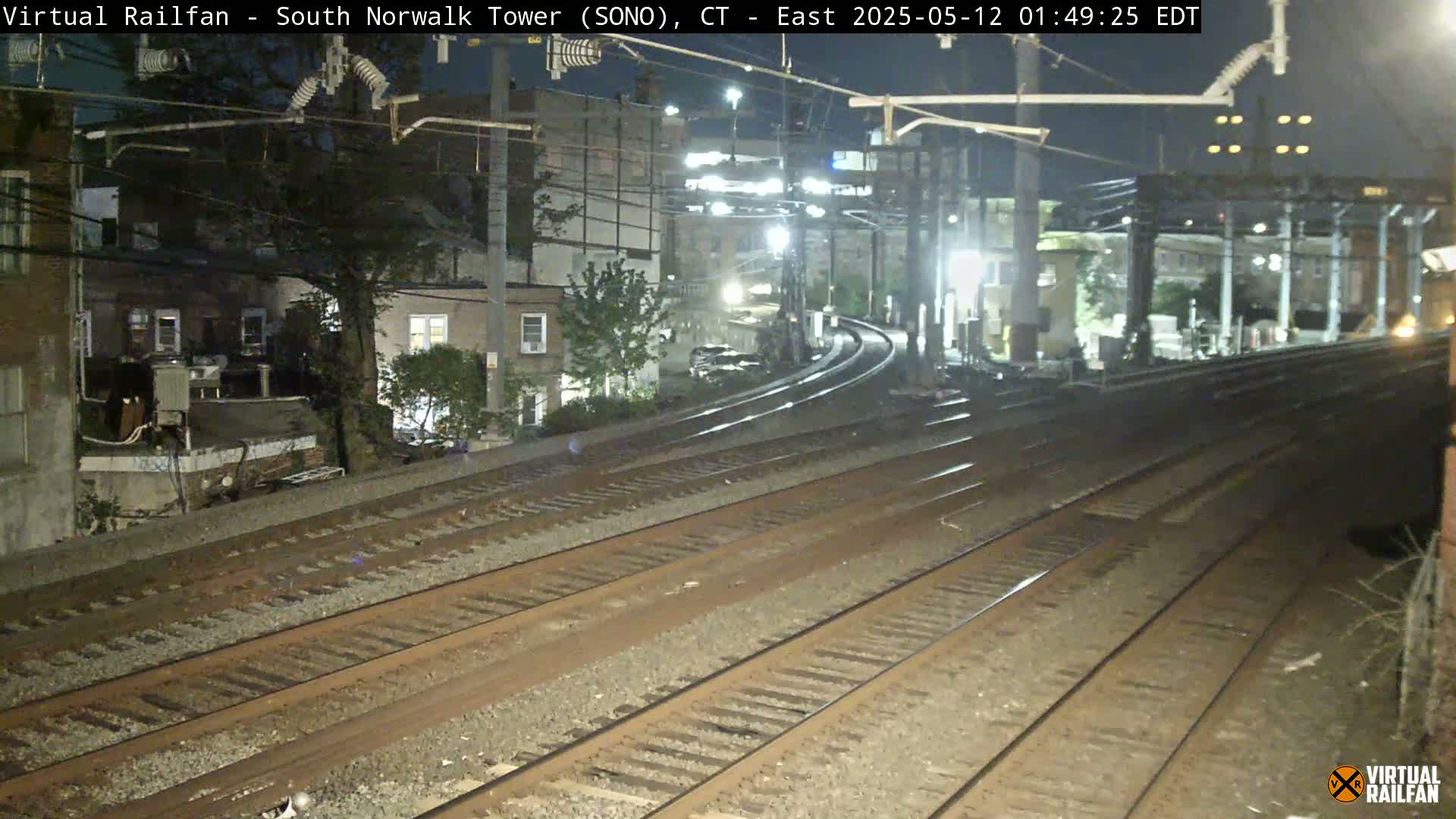 At night, several sets of train tracks curve away from a camera positioned near residential buildings under overhead electrical lines.